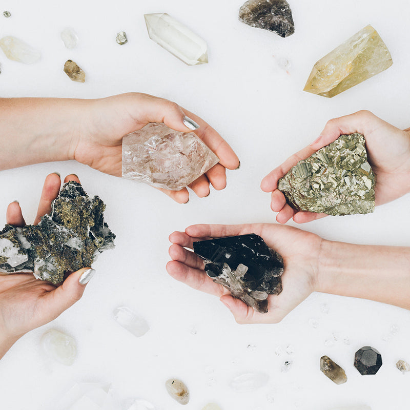 Flatlay of two women’s hands holding protective crystals including Pyrite, Smoky Quartz, and Clear Quartz, with other stones arranged for energy shielding.