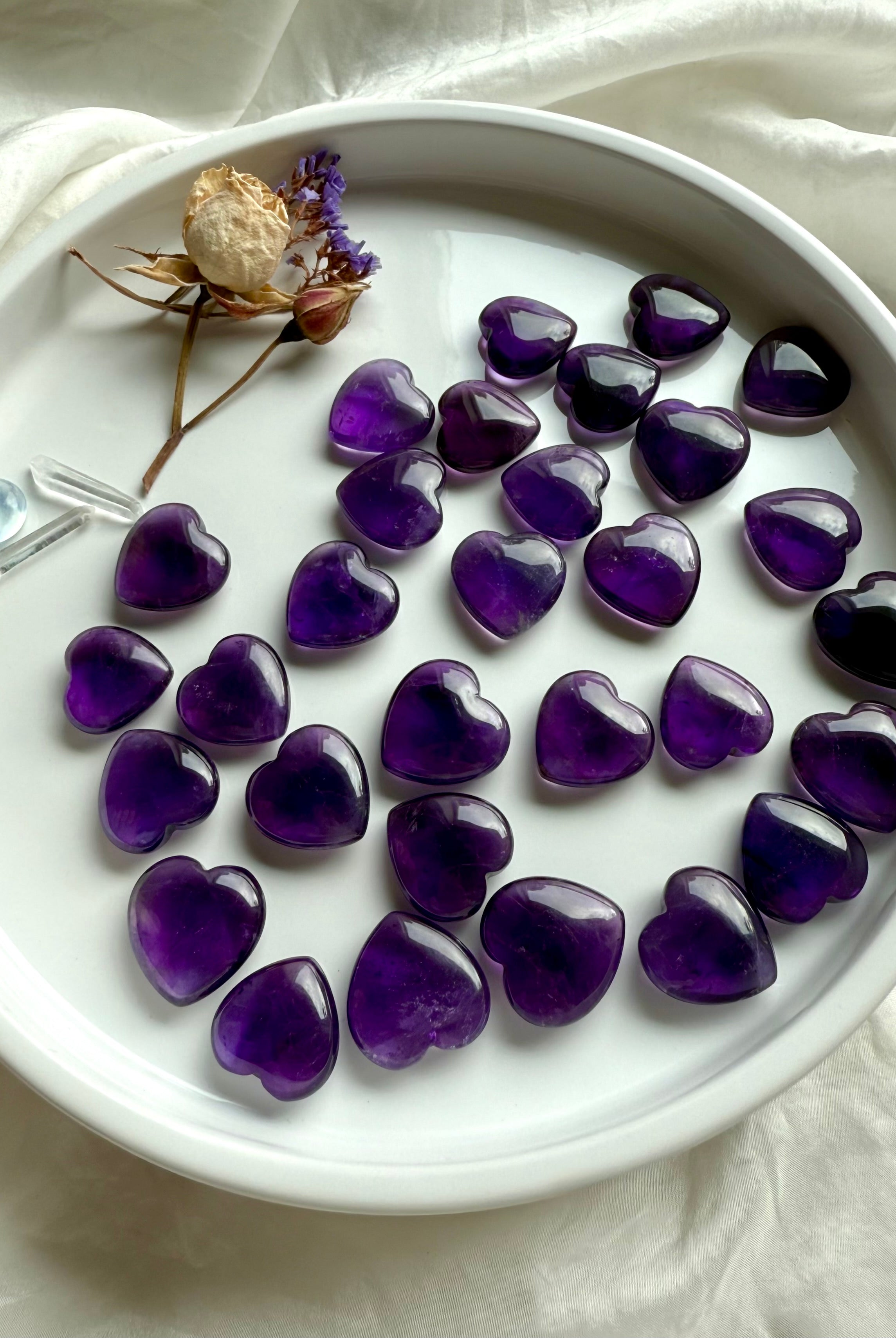 Assorted Amethyst crystal hearts arranged on a white dish with dried flowers and clear quartz — crown chakra healing stones for intuition and divine guidance.
