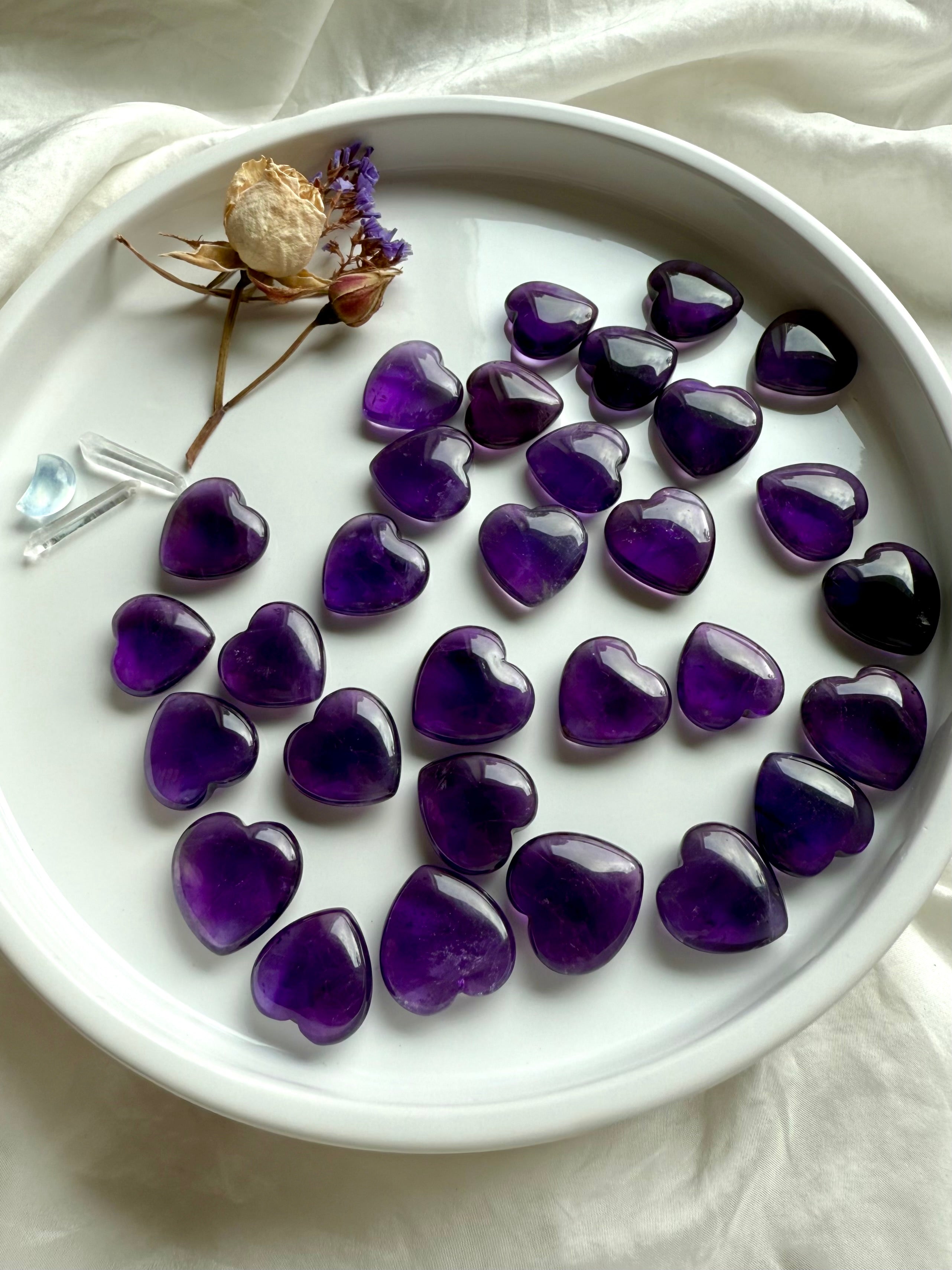Assorted Amethyst crystal hearts arranged on a white dish with dried flowers and clear quartz — crown chakra healing stones for intuition and divine guidance.