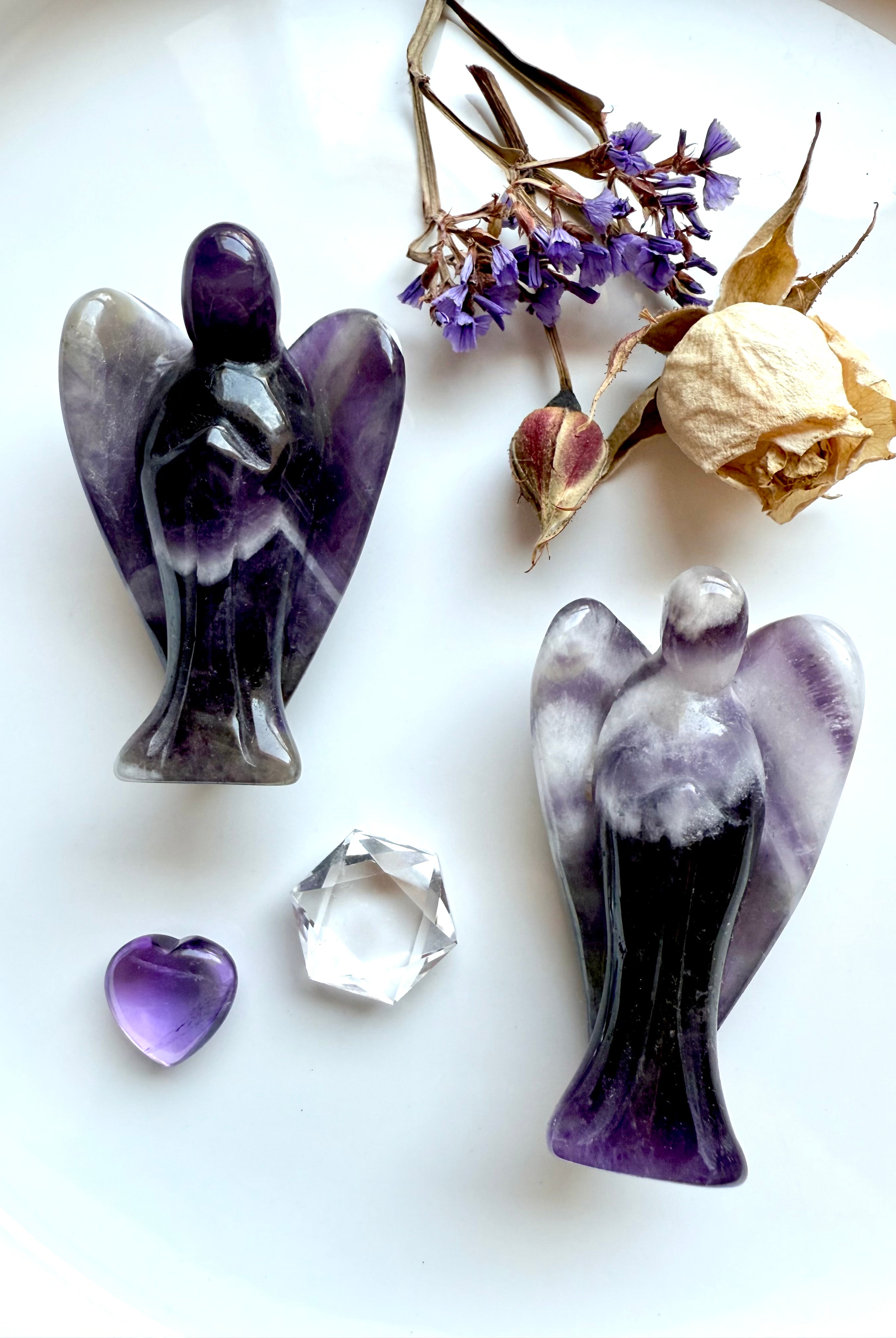 Dream Amethyst angel carvings with chevron banding, styled on a white plate with dried rose, purple statice flowers, Amethyst heart, and clear quartz gemstone – crystal angels for healing, grief, and spiritual protection