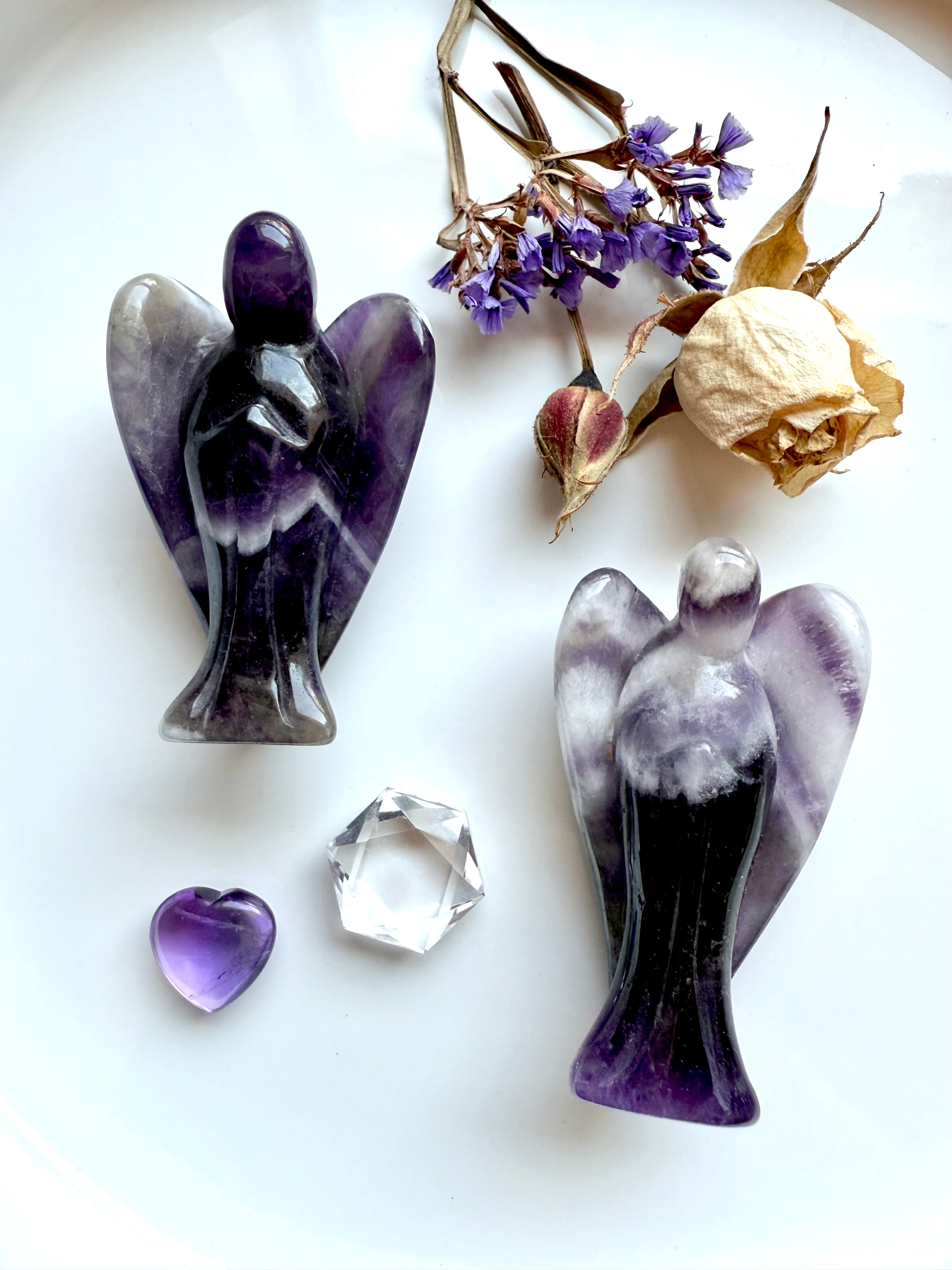Dream Amethyst angel carvings with chevron banding, styled on a white plate with dried rose, purple statice flowers, Amethyst heart, and clear quartz gemstone – crystal angels for healing, grief, and spiritual protection