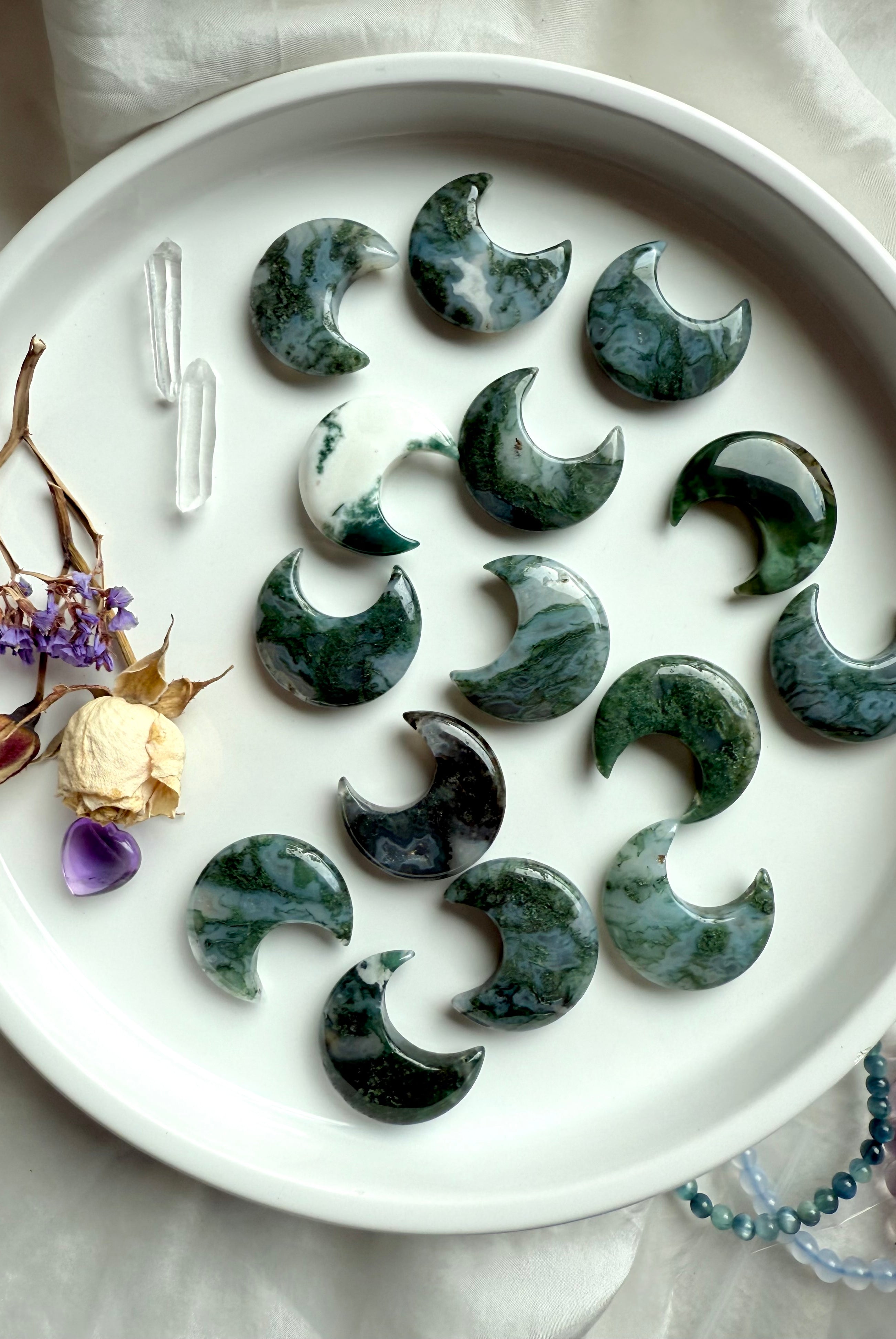 Moss Agate crescent moons with Clear Quartz points and dried flowers arranged on a white plate in a celestial altar flatlay.