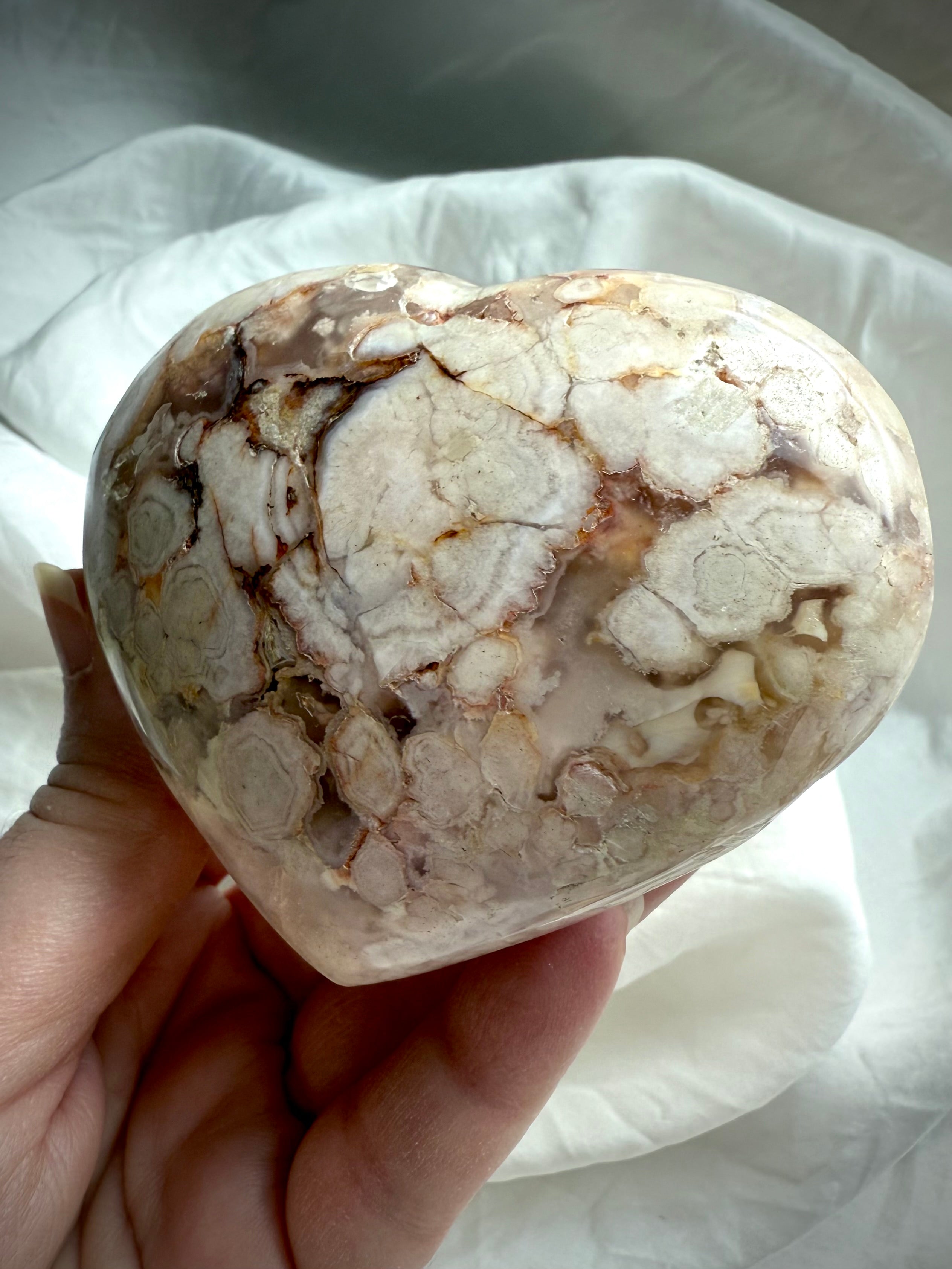 Flower Agate heart tilted in hand to show its natural blossom clusters and warm earthy tones โ supportive heart chakra crystal for healing and personal transformation.
