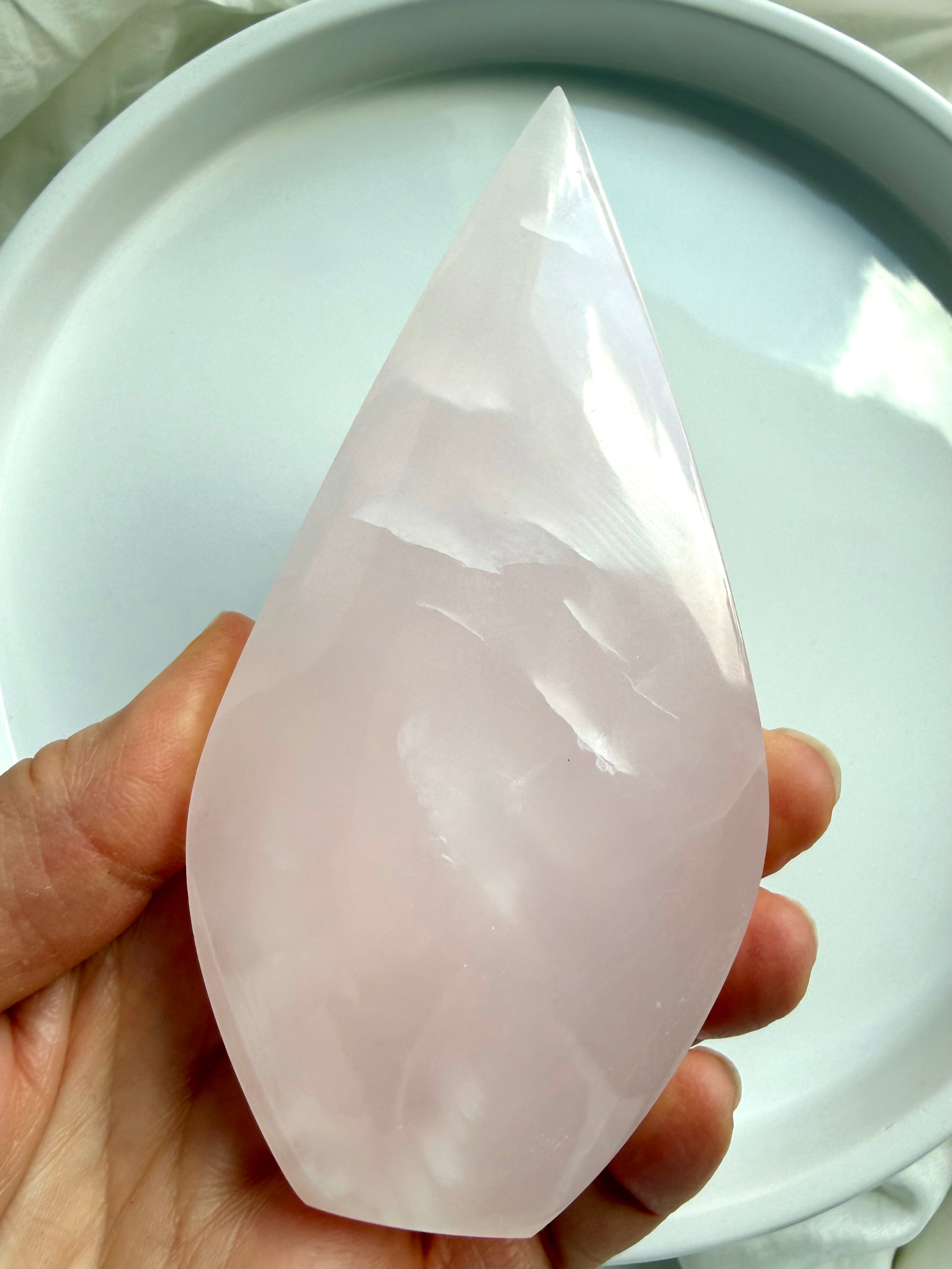 Mangano Calcite flame crystal held in hand for scale, featuring soft pink translucency and a smooth polished finish from Pakistan