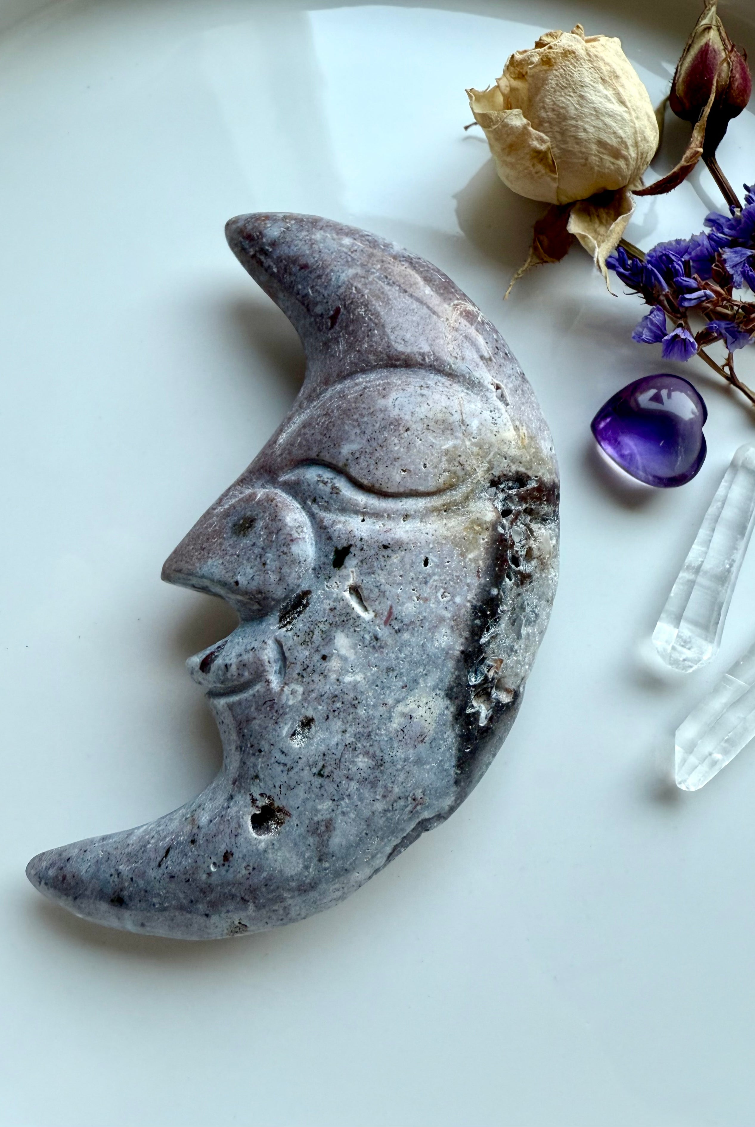 Purple Ocean Jasper moon face crystal carving displayed with quartz points, amethyst heart, and flowers.