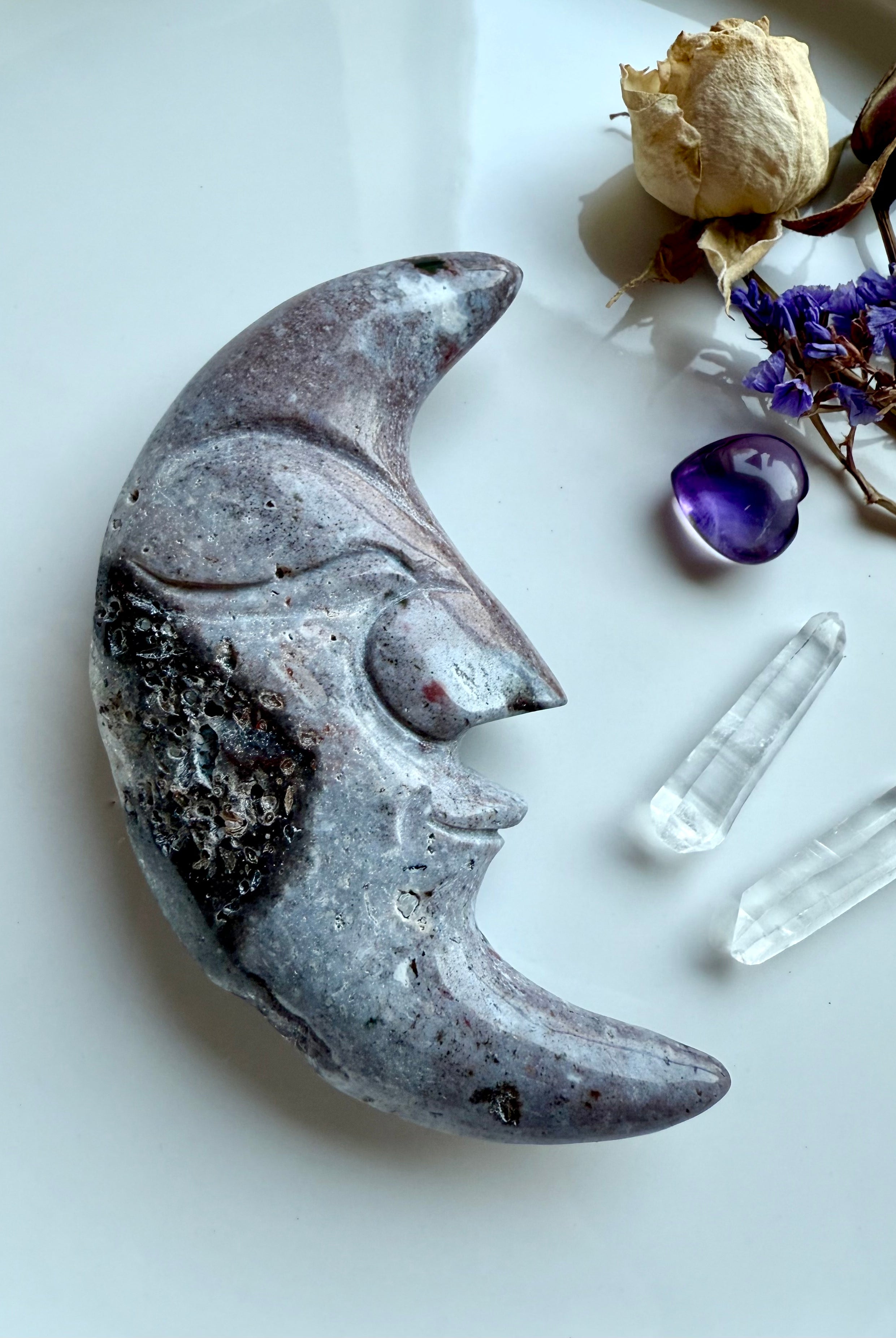 Purple Ocean Jasper moon face carving displayed with clear quartz points, amethyst heart, and flowers.