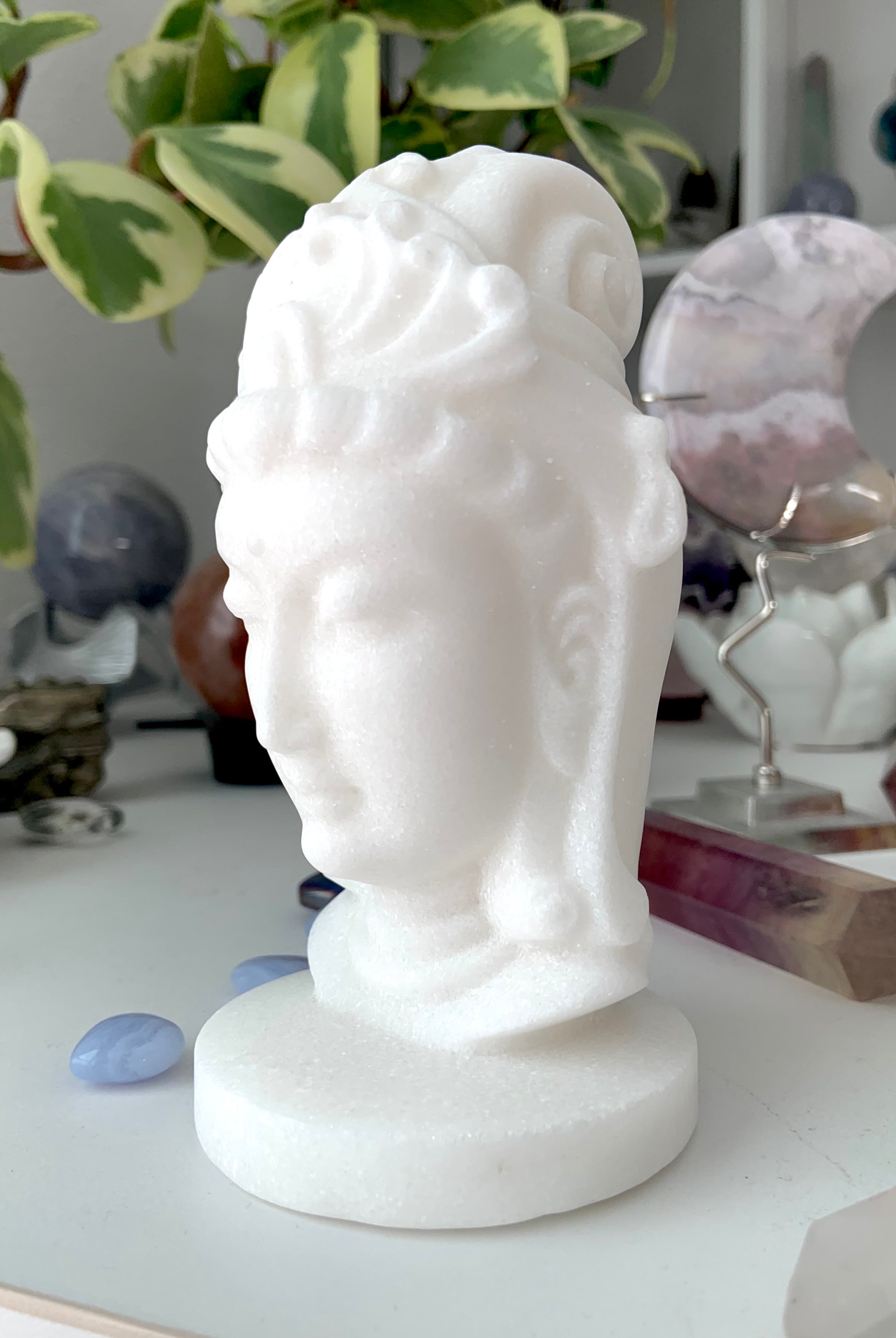 White Jade Quan Yin carving shown on a white table with other crystals and a plant in the background
