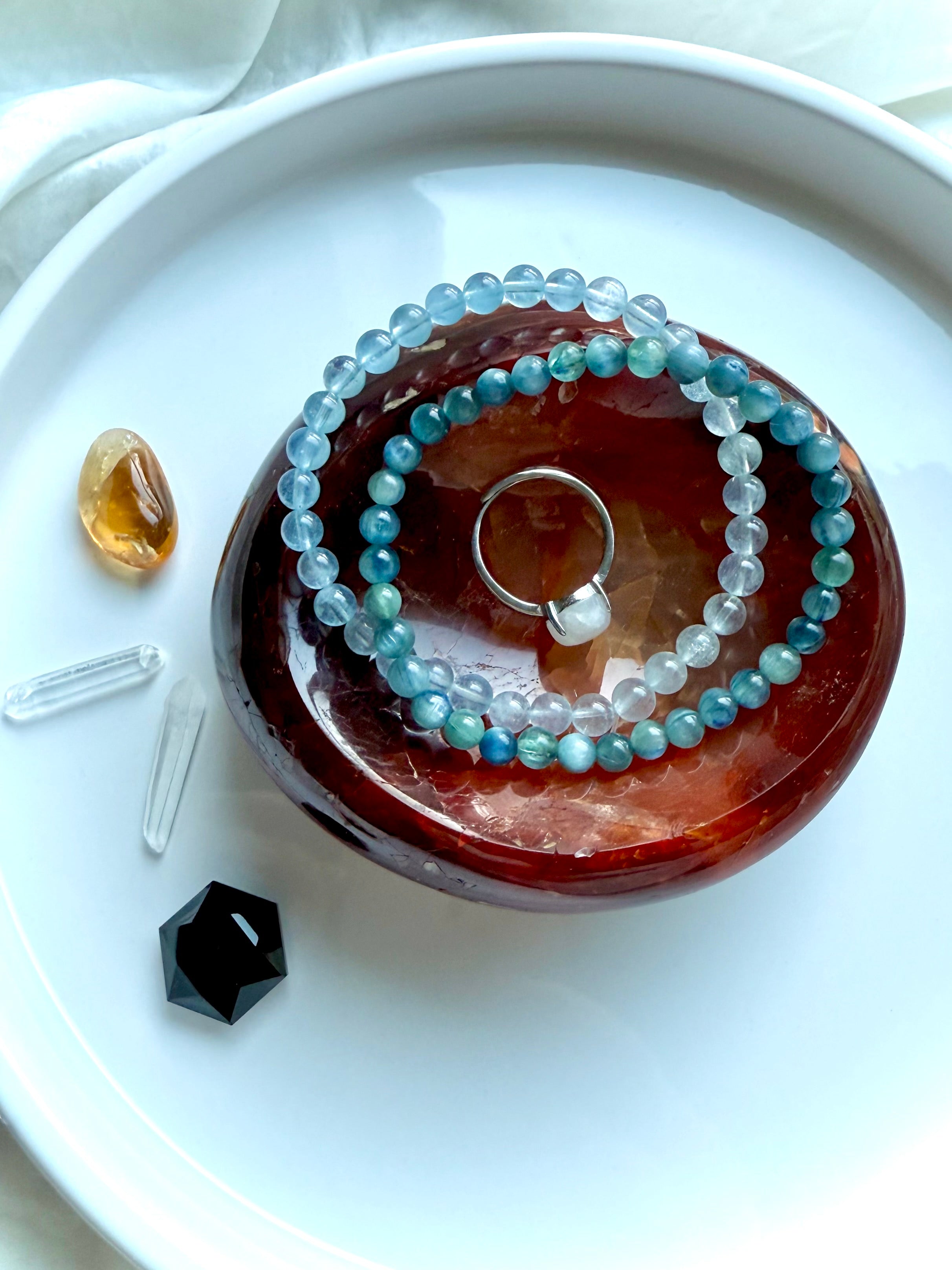 Polished Carnelian bowl holding aquamarine and kyanite bracelets with a moonstone ring, surrounded by healing crystals – chakra jewelry display