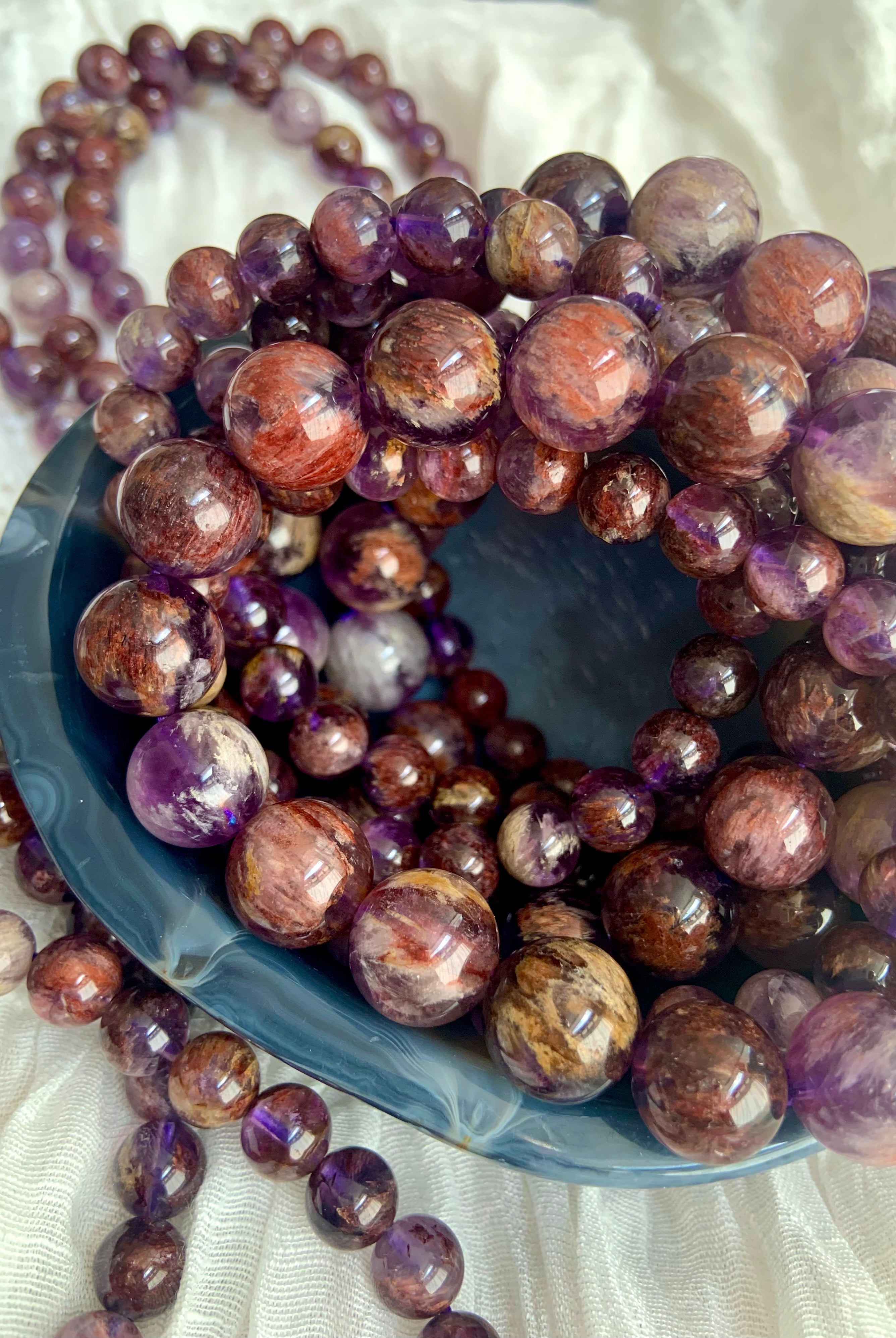 Super Seven crystal bracelets in varied bead sizes arranged in a bowl showing natural purple and mineral inclusions