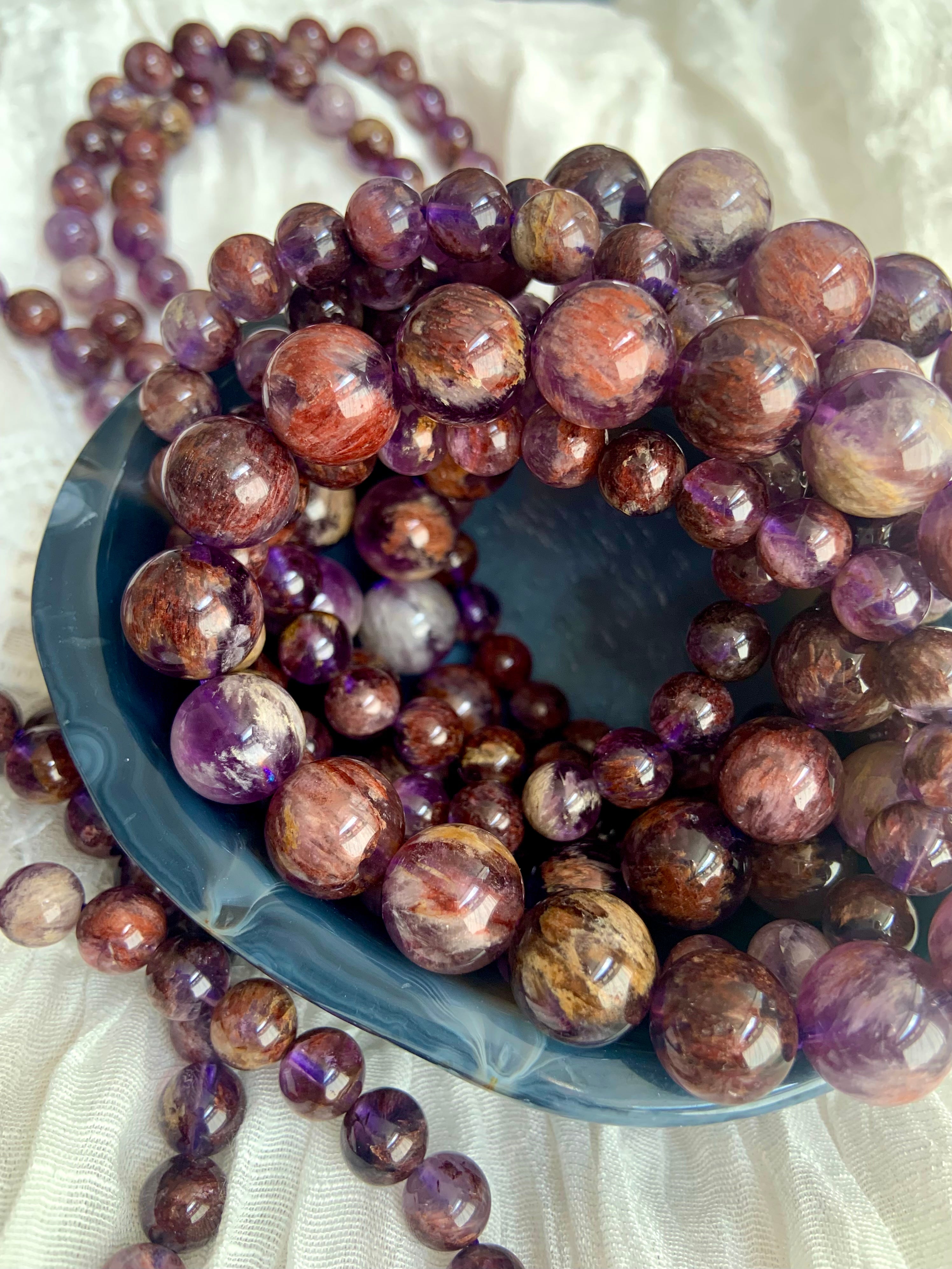 Super Seven crystal bracelets in varied bead sizes arranged in a bowl showing natural purple and mineral inclusions