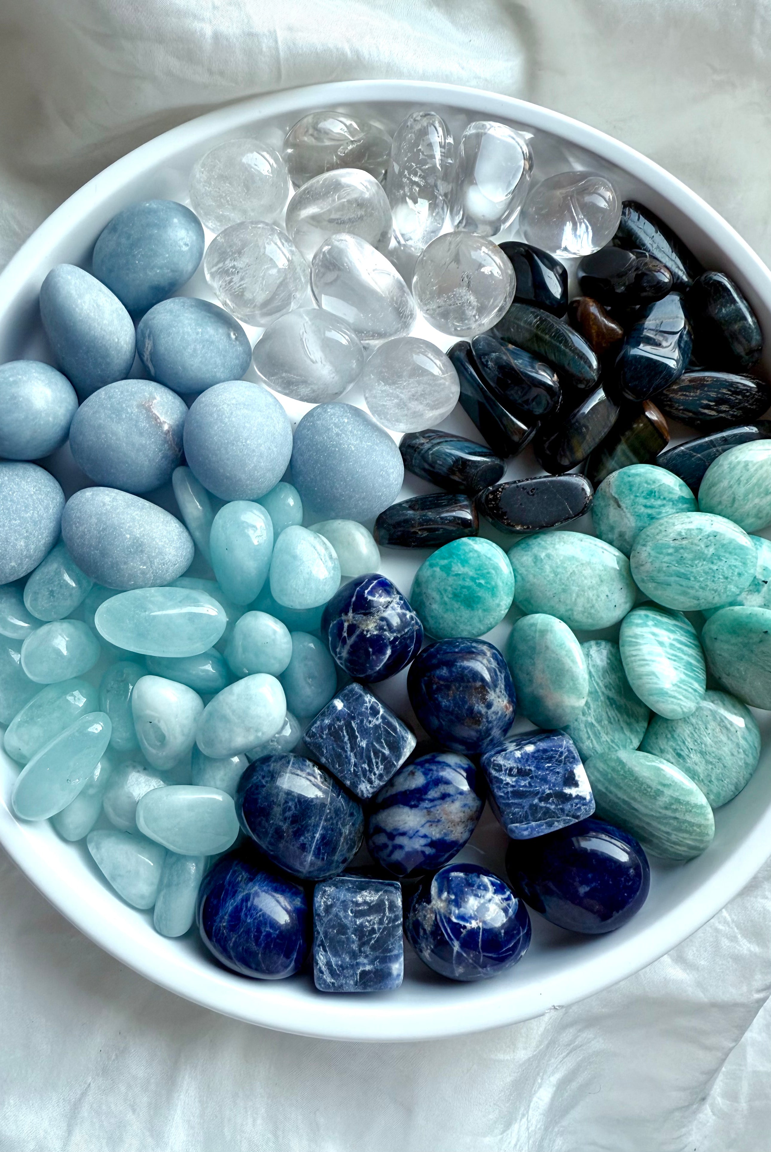 Bowl of throat chakra crystals — Angelite, Sodalite, Amazonite, Aquamarine, Blue Tiger’s Eye, and Clear Quartz healing stones