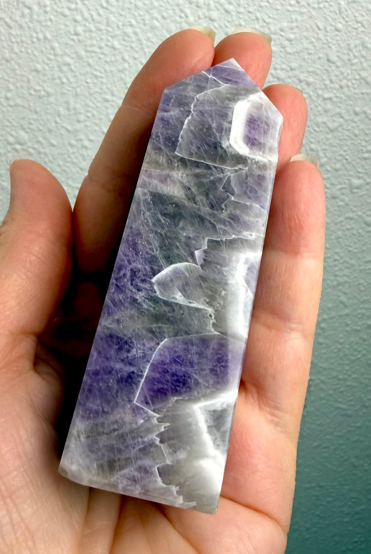 Polished Dream Amethyst crystal tower held in hand, showing its size, lavender purple coloring and distinctive white quartz chevrons