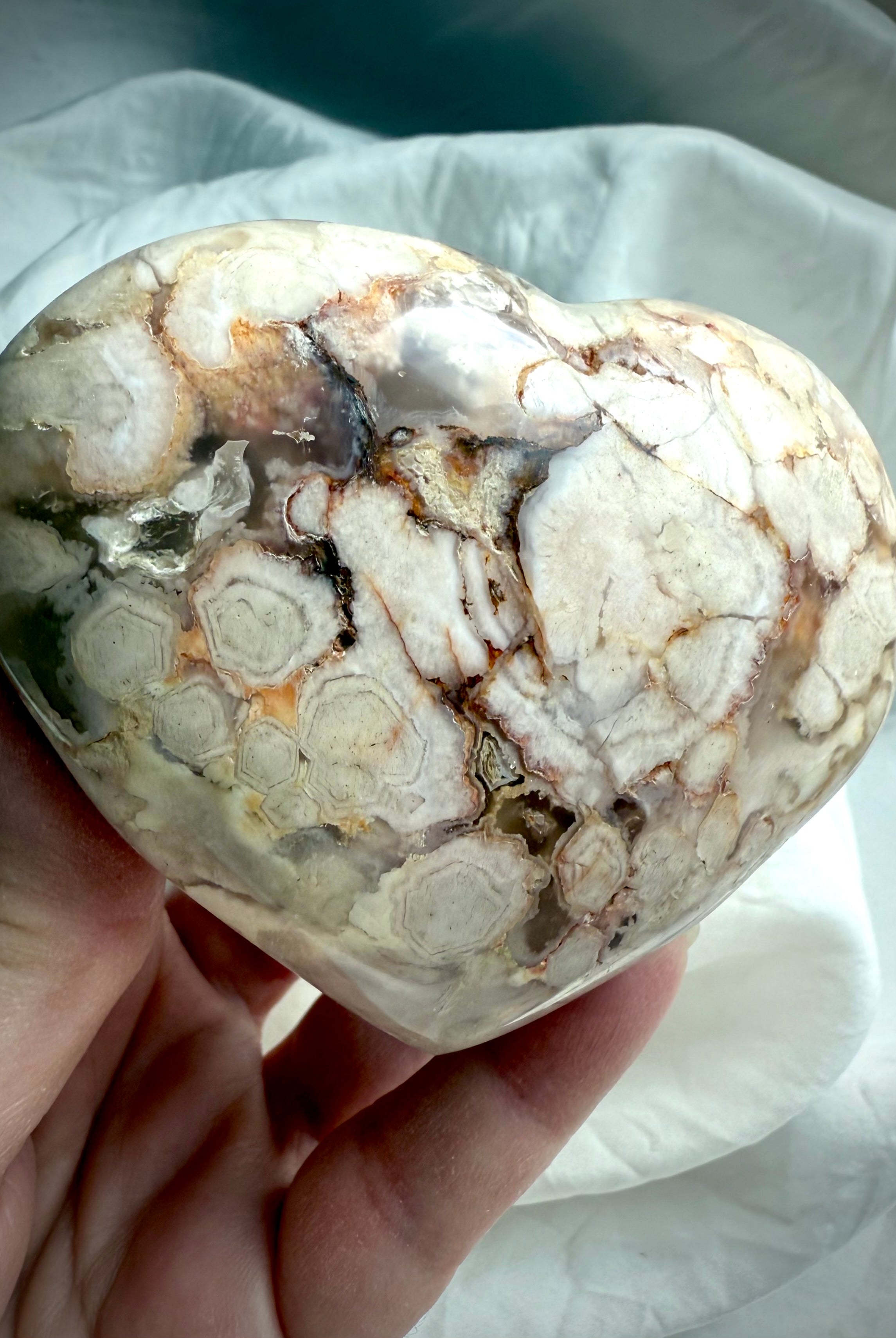 Hand holding a Flower Agate heart at an angle, capturing its blooming patterns and soft pastel hues — gentle crystal for emotional balance, resilience, and renewal.