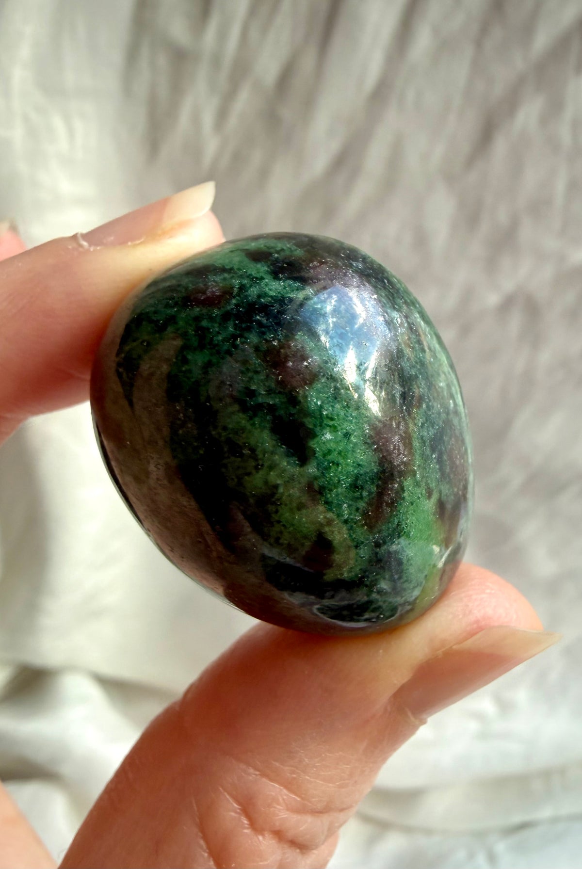 Natural Ruby in Zoisite tumbled stone held between fingers, showing rich emerald green, dark mineral inclusions and ruby pink coloring