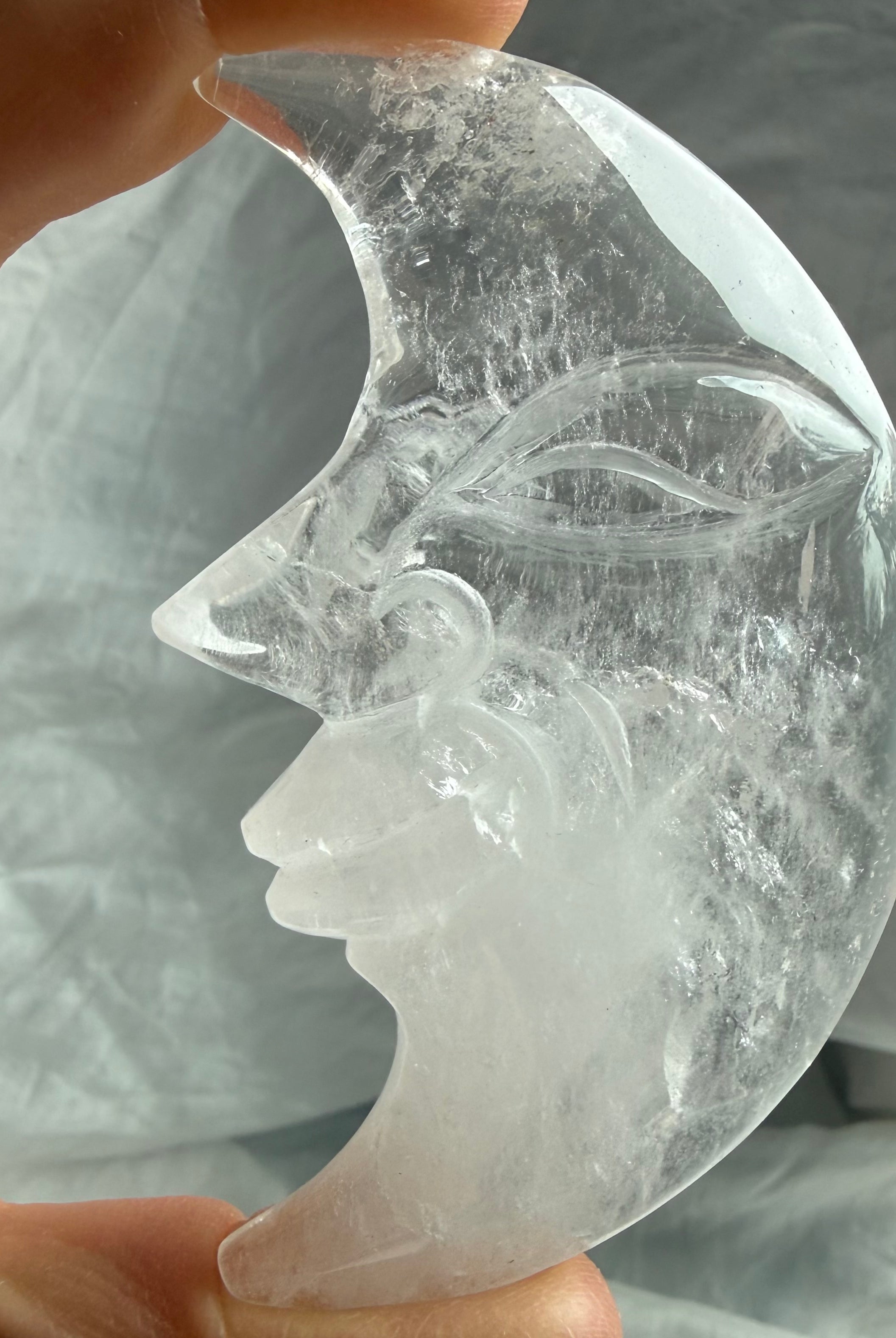 Clear Quartz Moon Face carving held in natural light, revealing exceptional clarity, smooth glossy polish, and celestial design