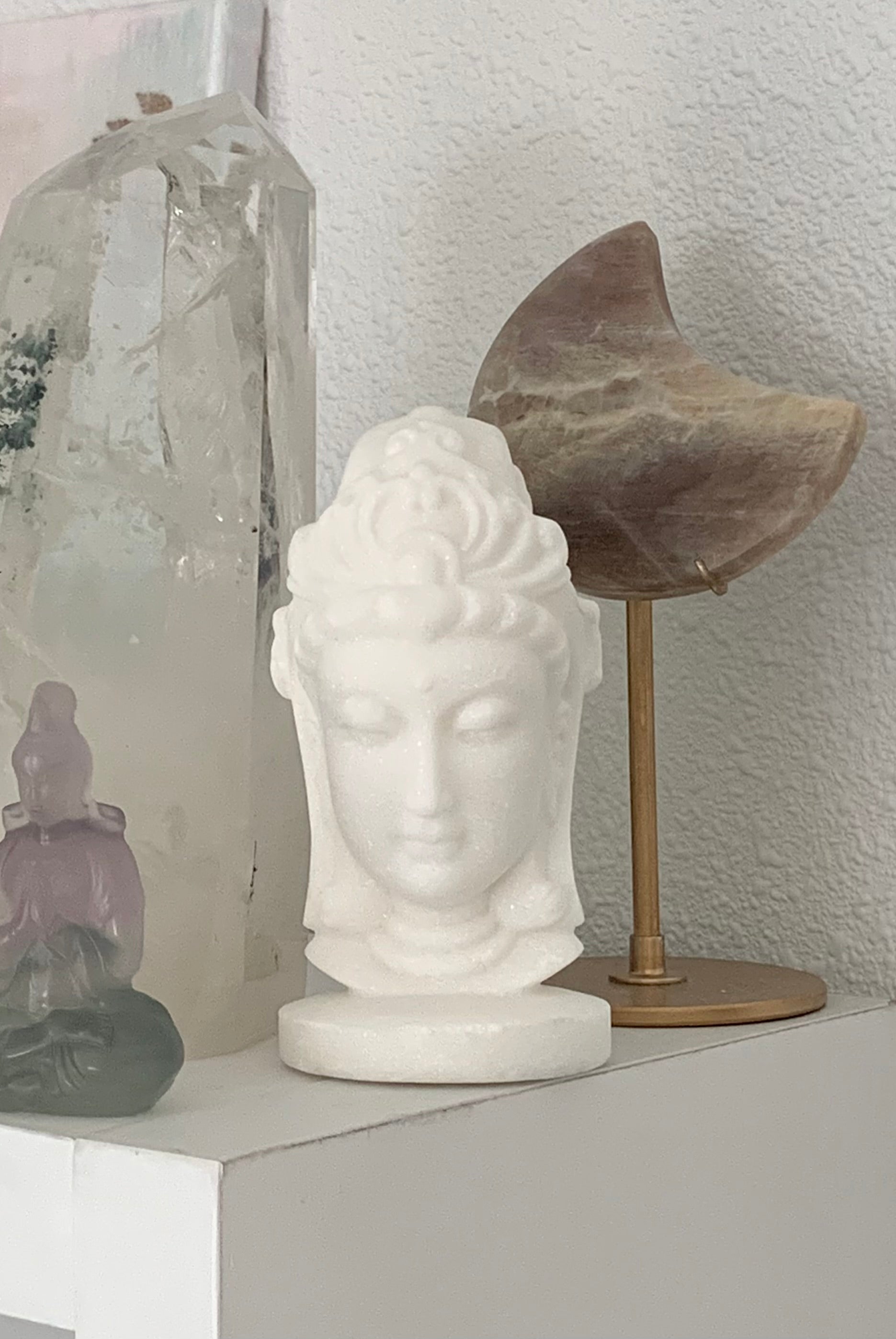 White Jade Quan Yin Statue shown on a shelf with some larger crystals and another Quan Yin figurine in the picture.