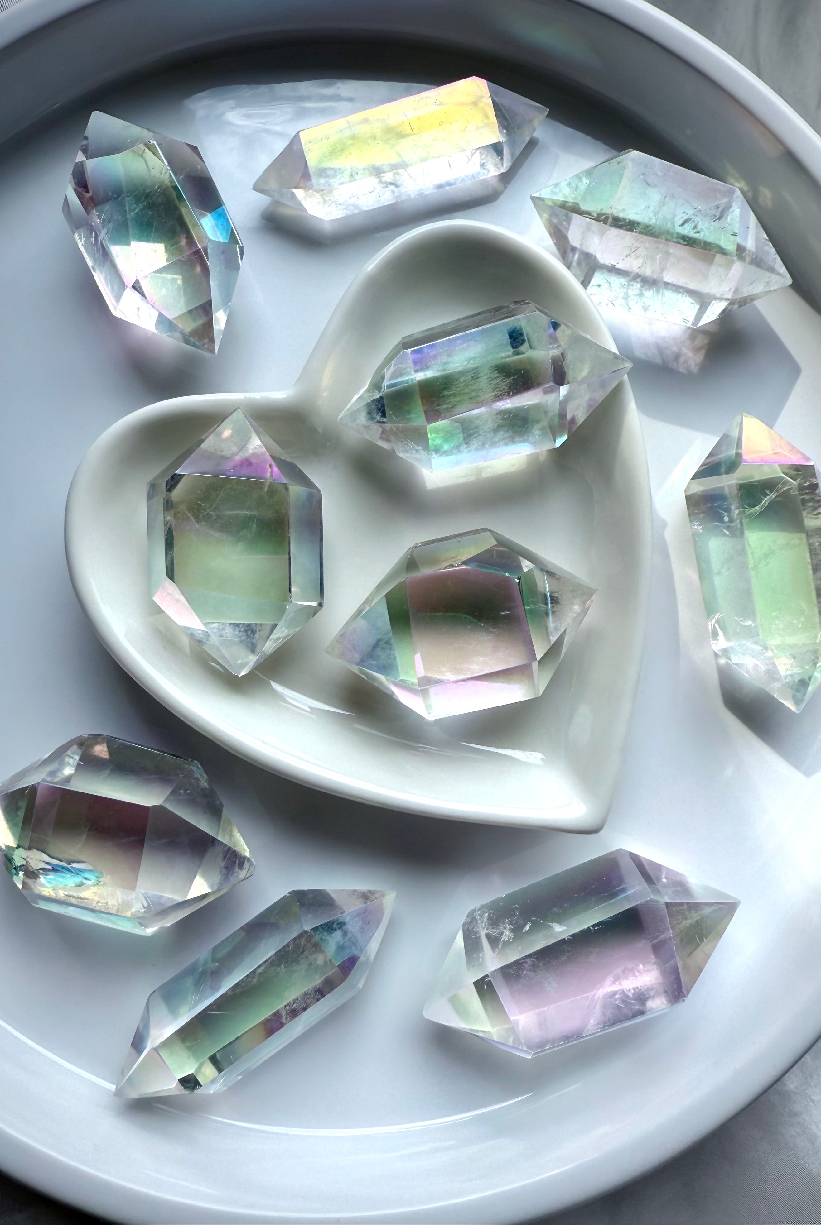 Group of Angel Aura Quartz DT crystals with iridescent rainbow aura coating, arranged in a heart bowl and white dish