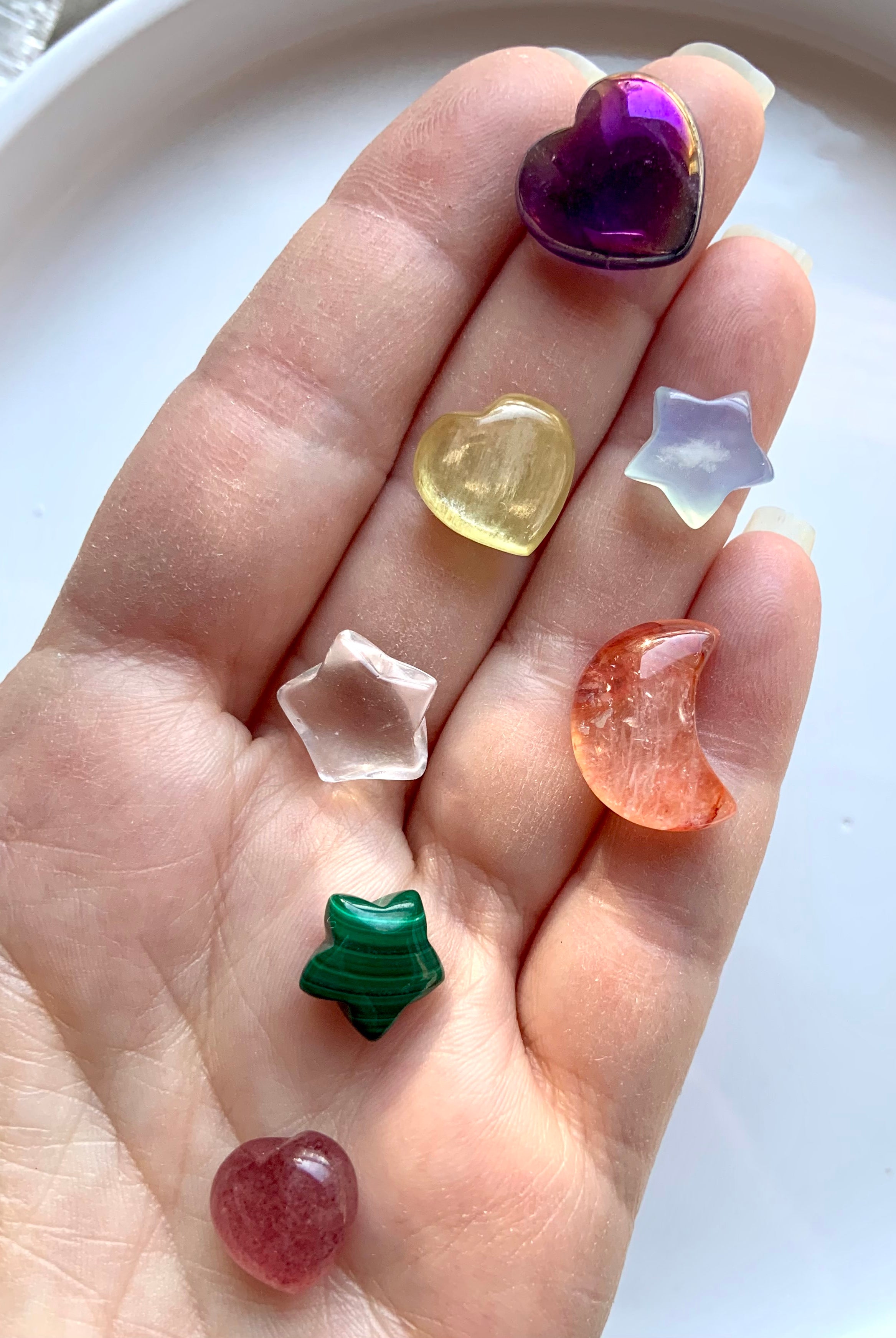 Hand holding 7 carved chakra crystals in heart, star & moon shapes in rainbow colors for each chakra