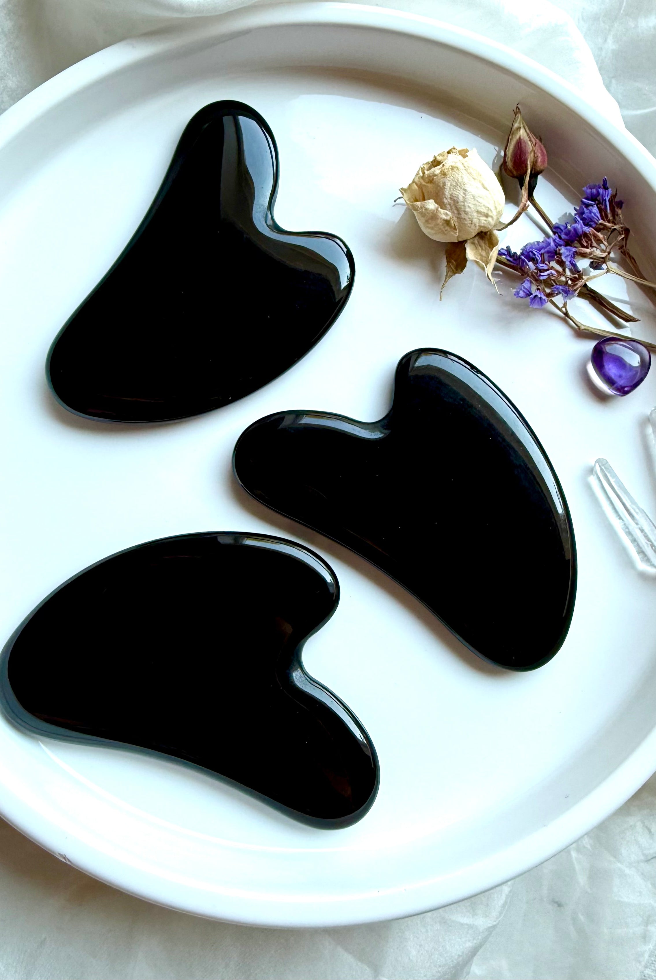 Three Black Obsidian crystal gua sha facial massage tools on white plate with dried flowers and amethyst accent