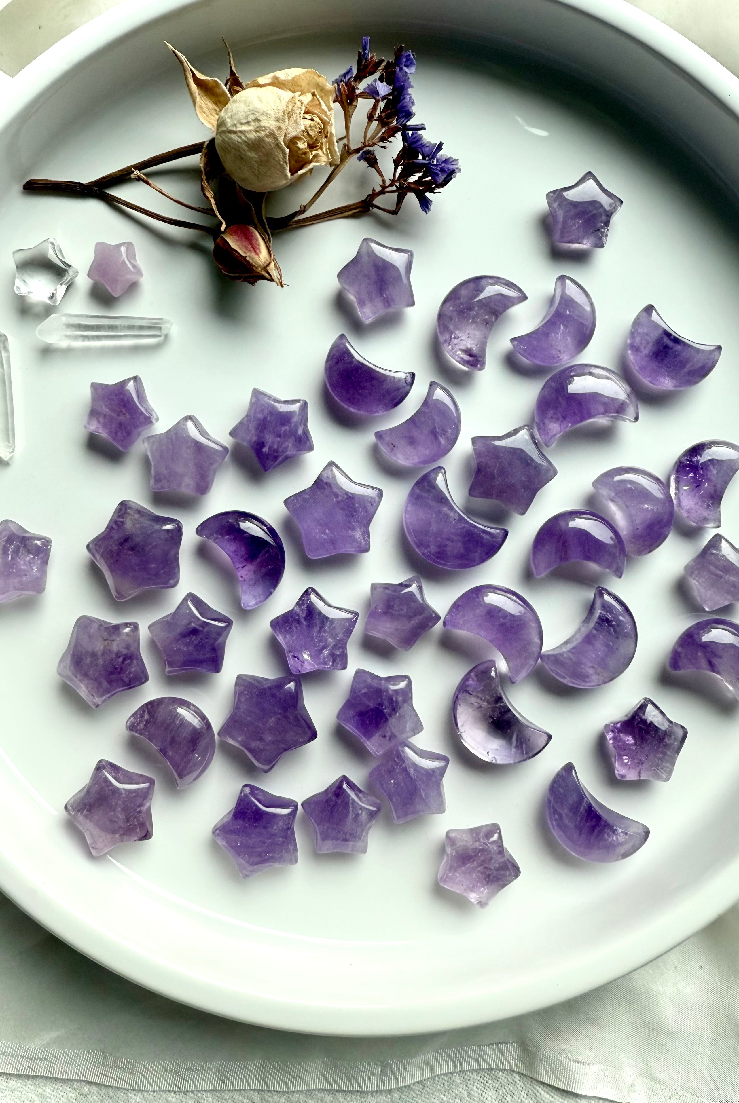 Mini amethyst moon and star carvings in soft violet hues arranged on a white ceramic tray with crystals and dried flowers