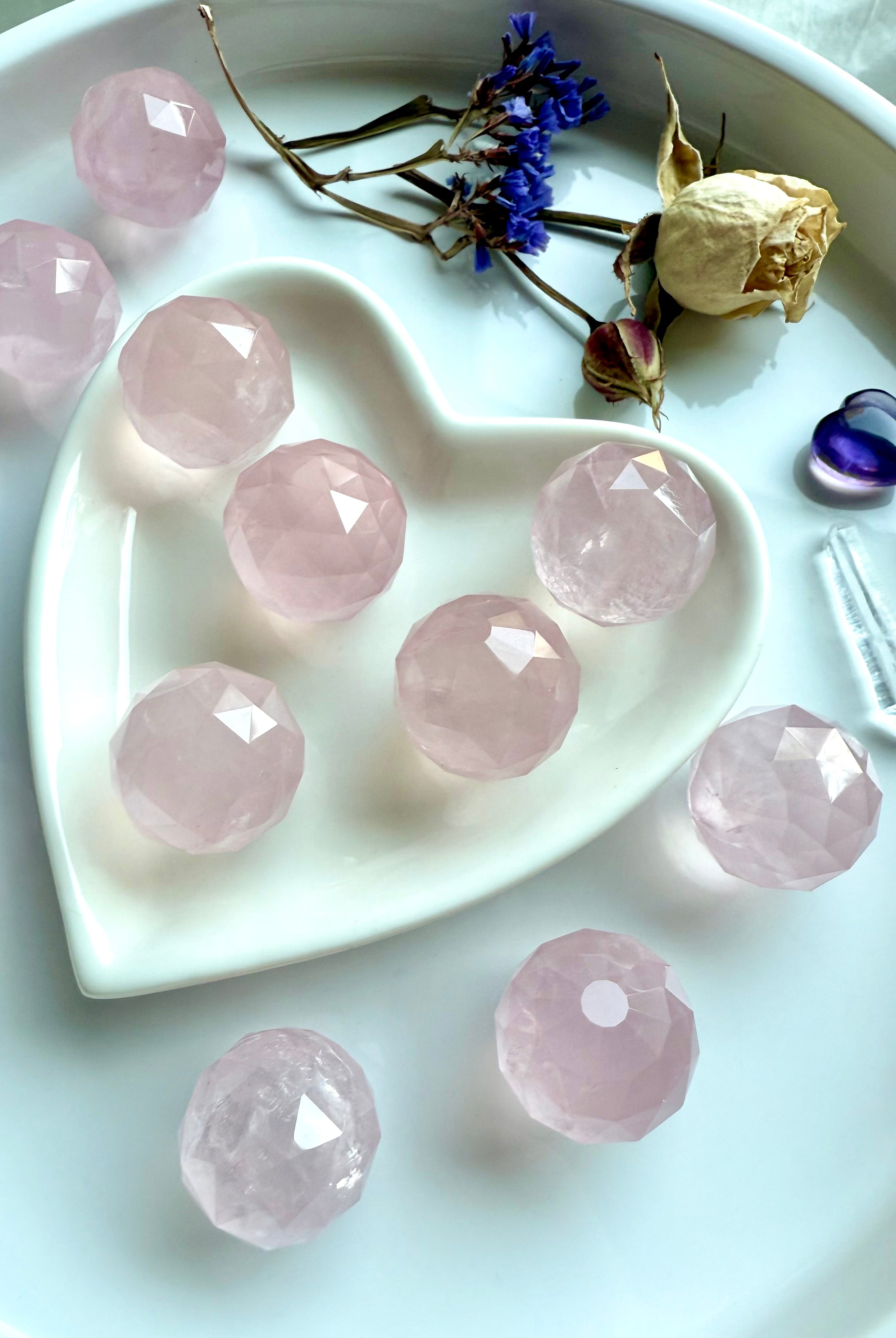 Mini faceted rose quartz crystal spheres on a heart-shaped white dish with dried flowers and crystals. Dreamy pink stones for heart chakra healing.