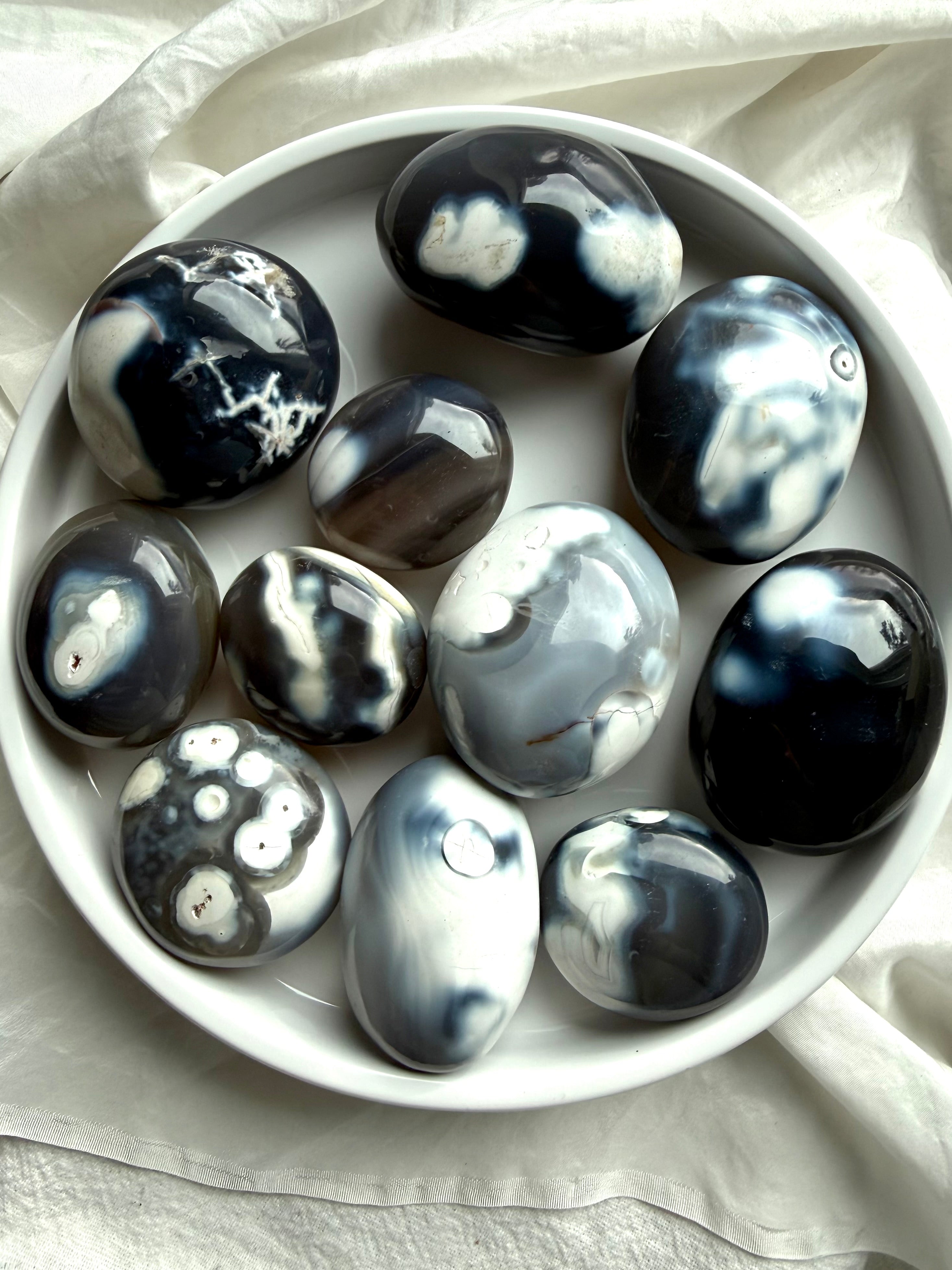 Orca Agate palm stones arranged in a white bowl, showing natural black, gray, and white banding for emotional grounding and calm