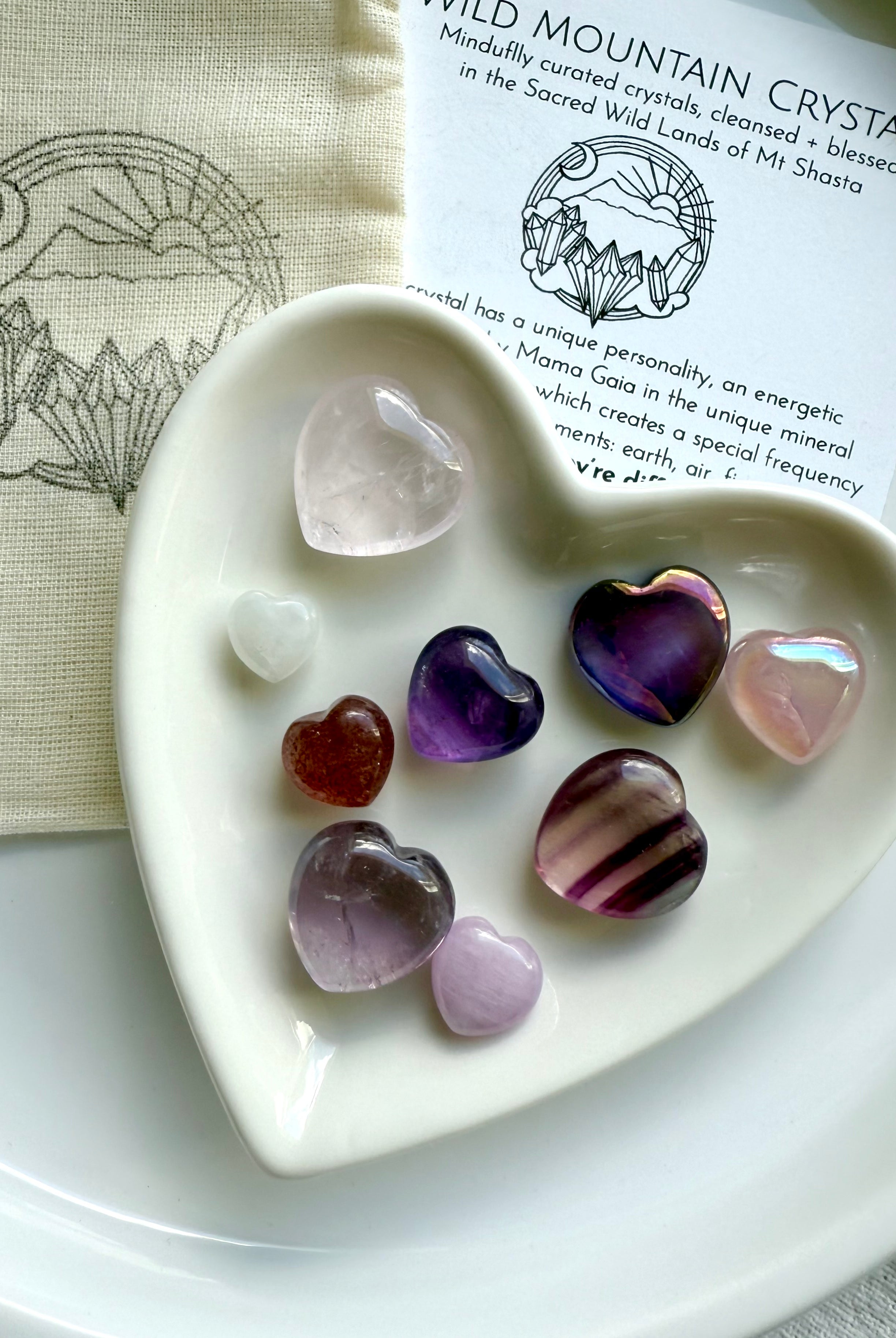 White heart-shaped bowl filled with nine crystal hearts, resting on cotton pouch and crystal info card, displayed on white plate.