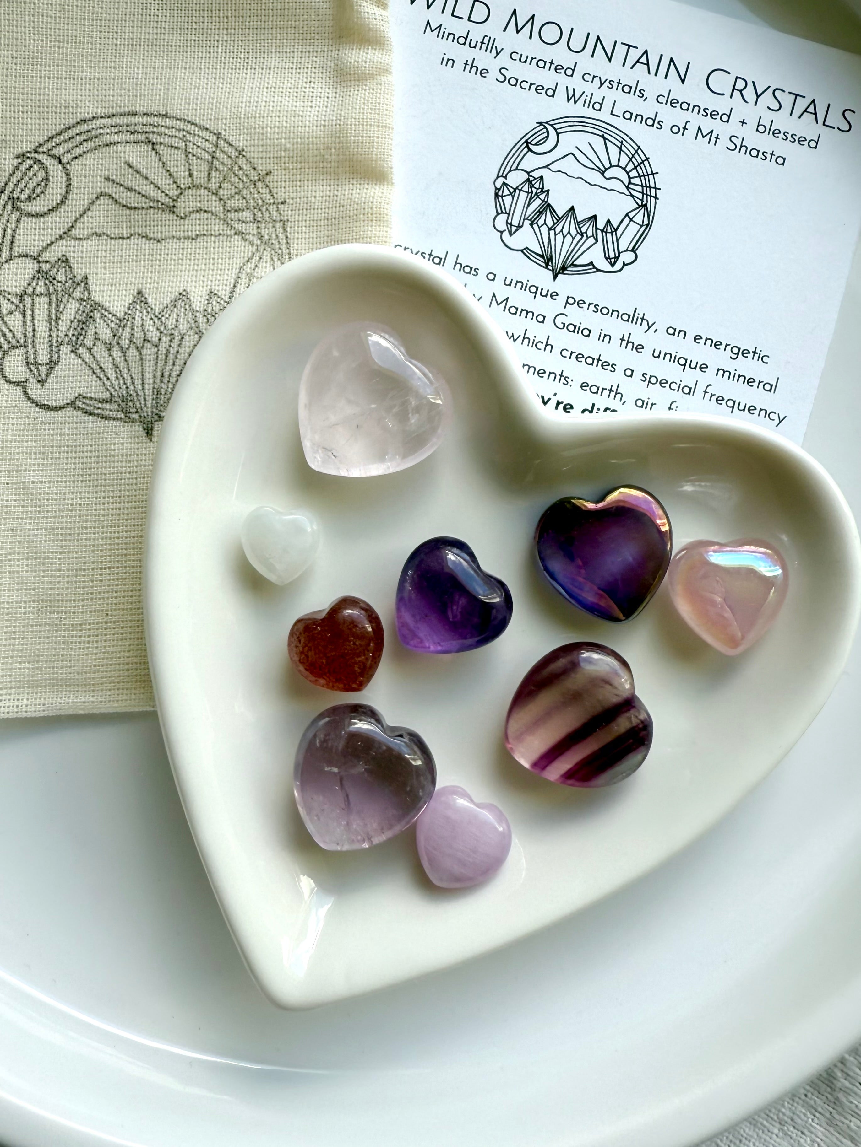 White heart-shaped bowl filled with nine crystal hearts, resting on cotton pouch and crystal info card, displayed on white plate.