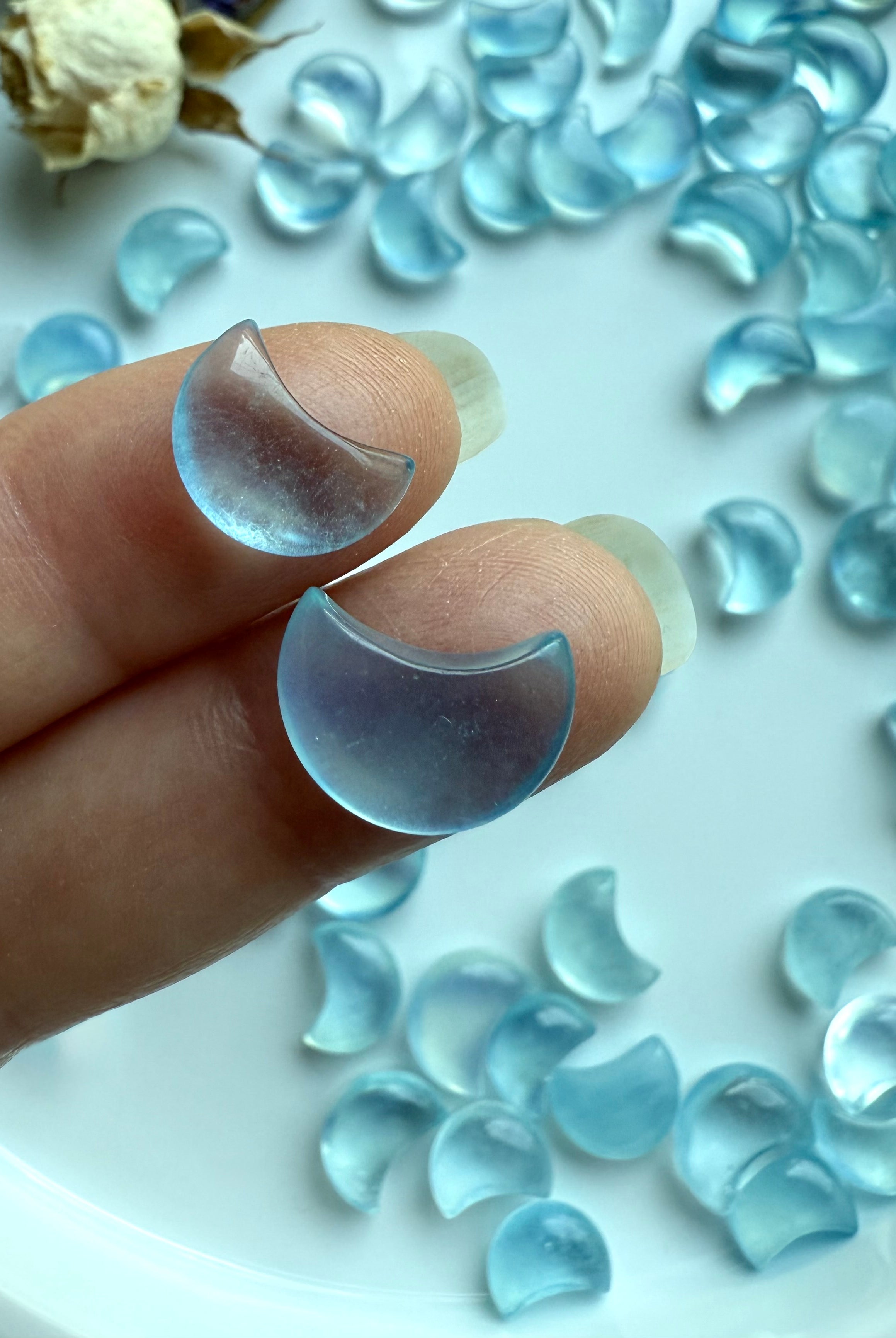 Closeup of two tiny Aquamarine crescent moon carvings resting on fingers, with a background of mini moons and dried flowers in soft lighting