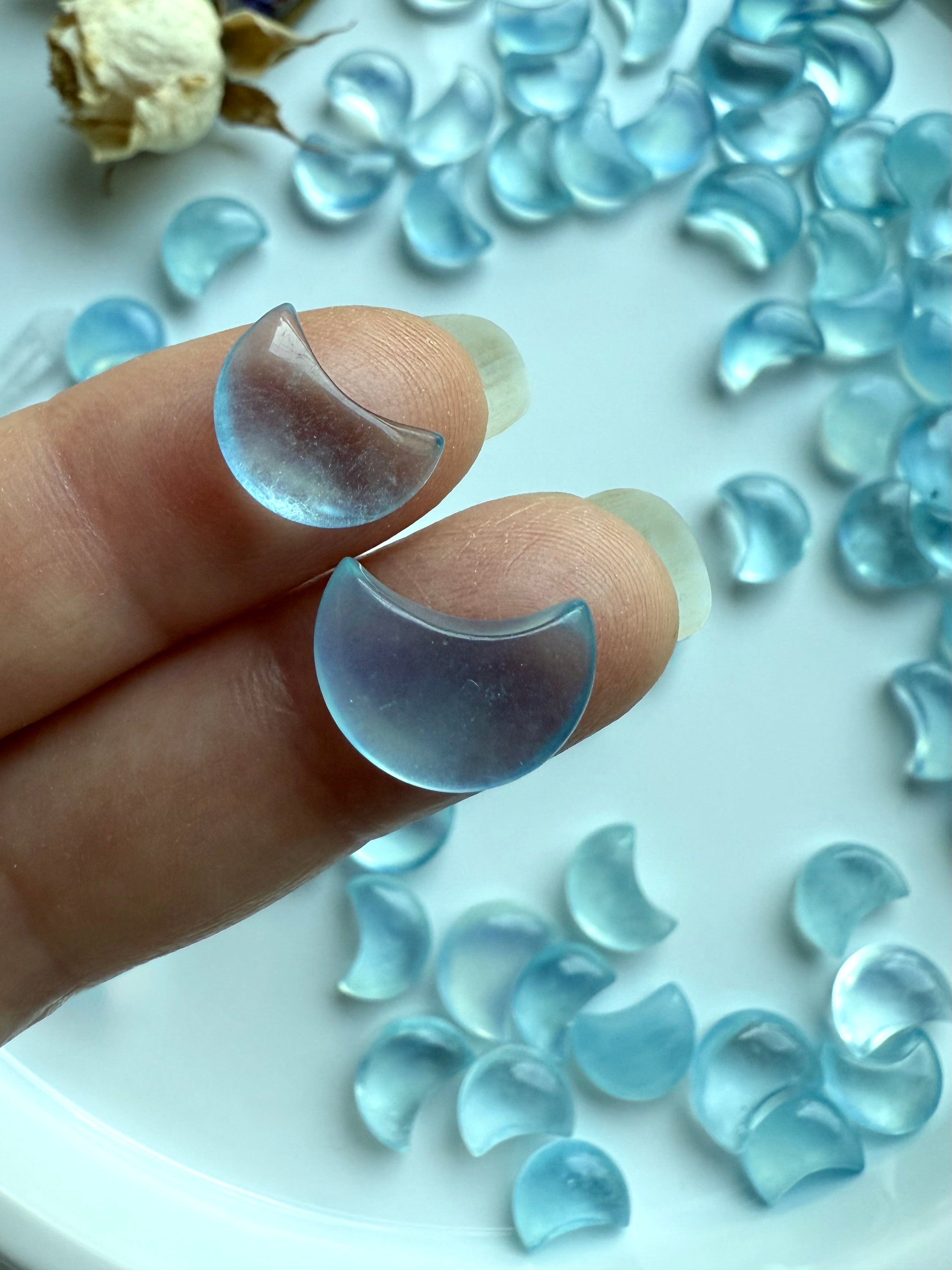 Closeup of two tiny Aquamarine crescent moon carvings resting on fingers, with a background of mini moons and dried flowers in soft lighting
