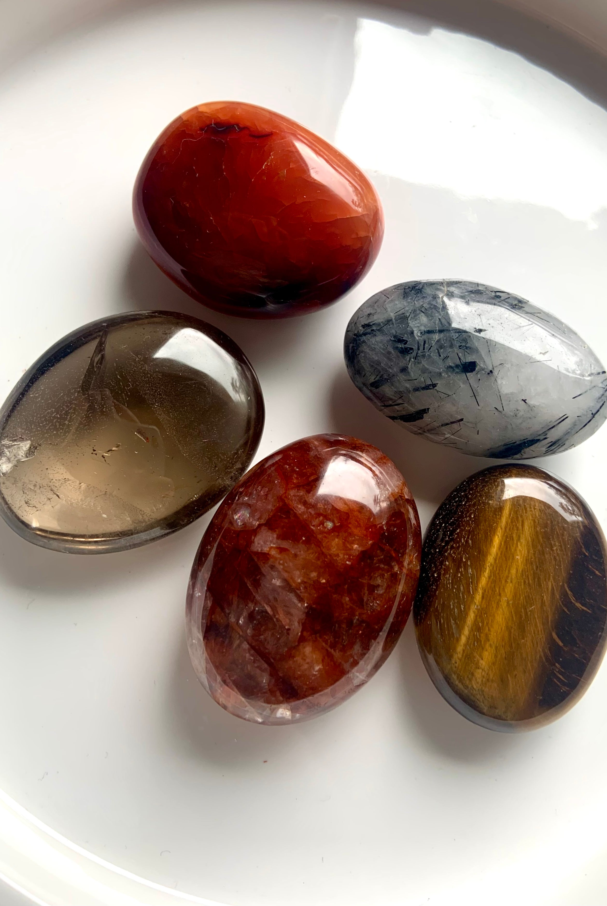 Closeup of five palm stones on white plate — includes Smokey Quartz, Tigers Eye, Carnelian, Fire Quartz & Tourmaline in Quartz for chakra healing.
