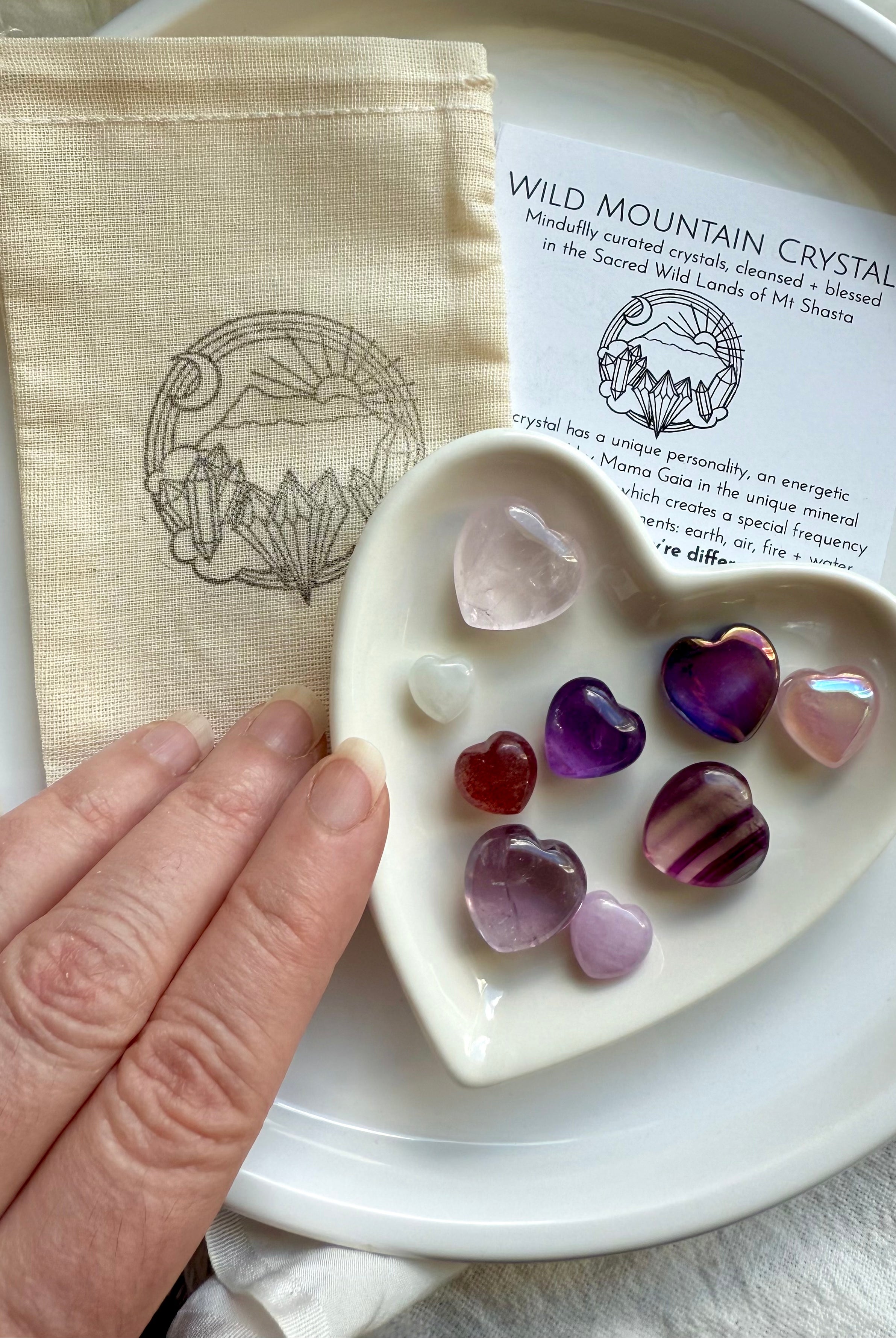 White heart-shaped bowl of crystal hearts on cotton pouch and info card, on a white plate, with hand reaching in.