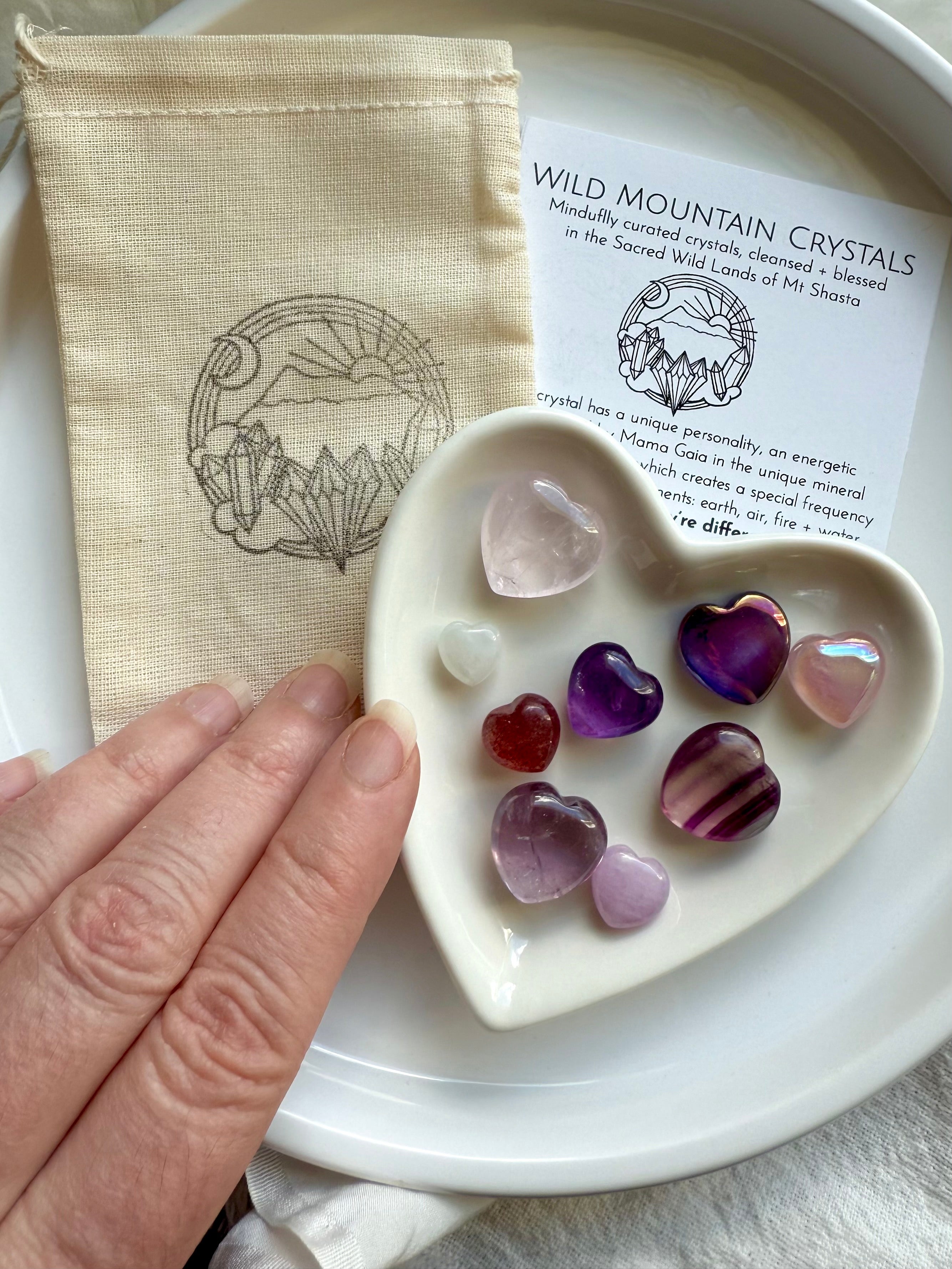 White heart-shaped bowl of crystal hearts on cotton pouch and info card, on a white plate, with hand reaching in.