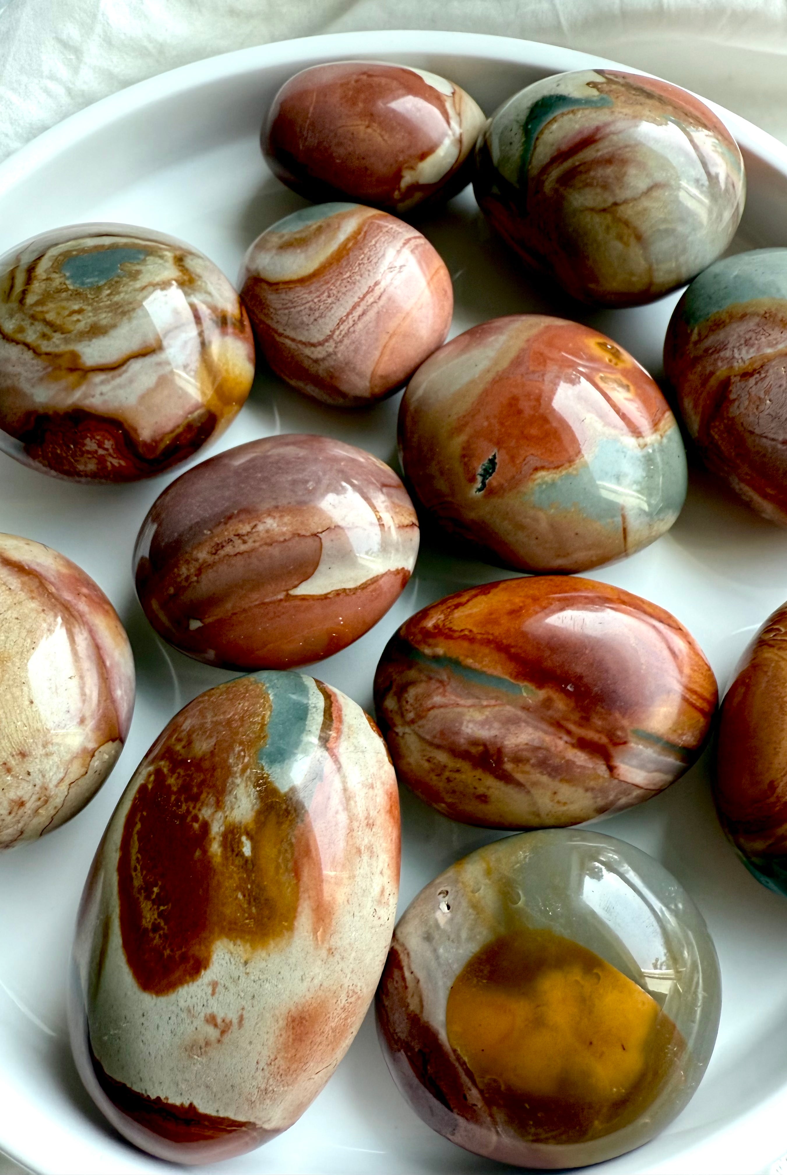 Polychrome Jasper palm stones on a white plate, showing earthy tones & swirled patterns for grounding energy