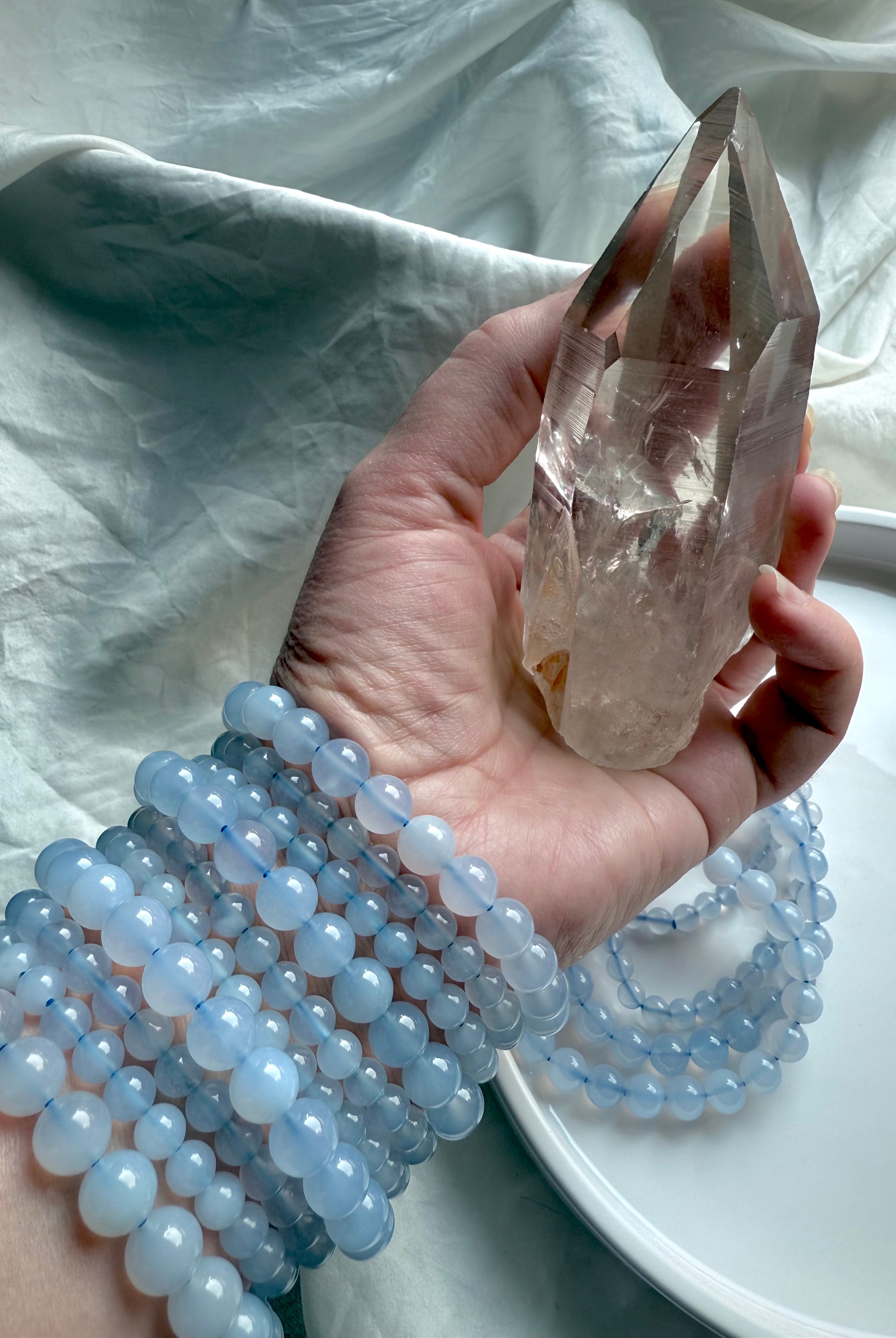 Hand holding a smoky quartz point with a stack of Blue Chalcedony bracelets — throat chakra healing stones with gentle calming energy.