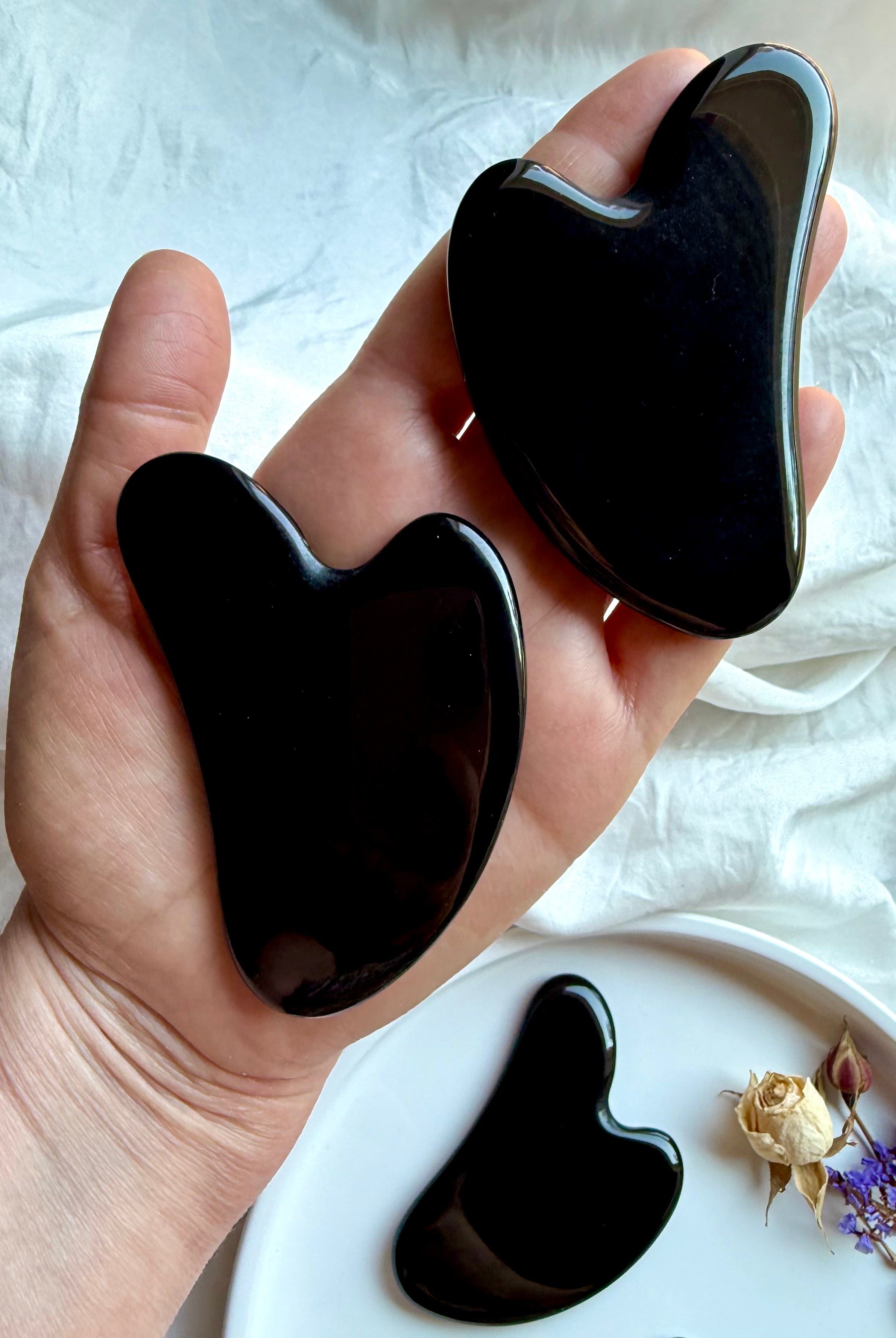 Hand holding two Black Obsidian crystal gua sha facial massage tools with curved contour shape, extra gua sha on plate below