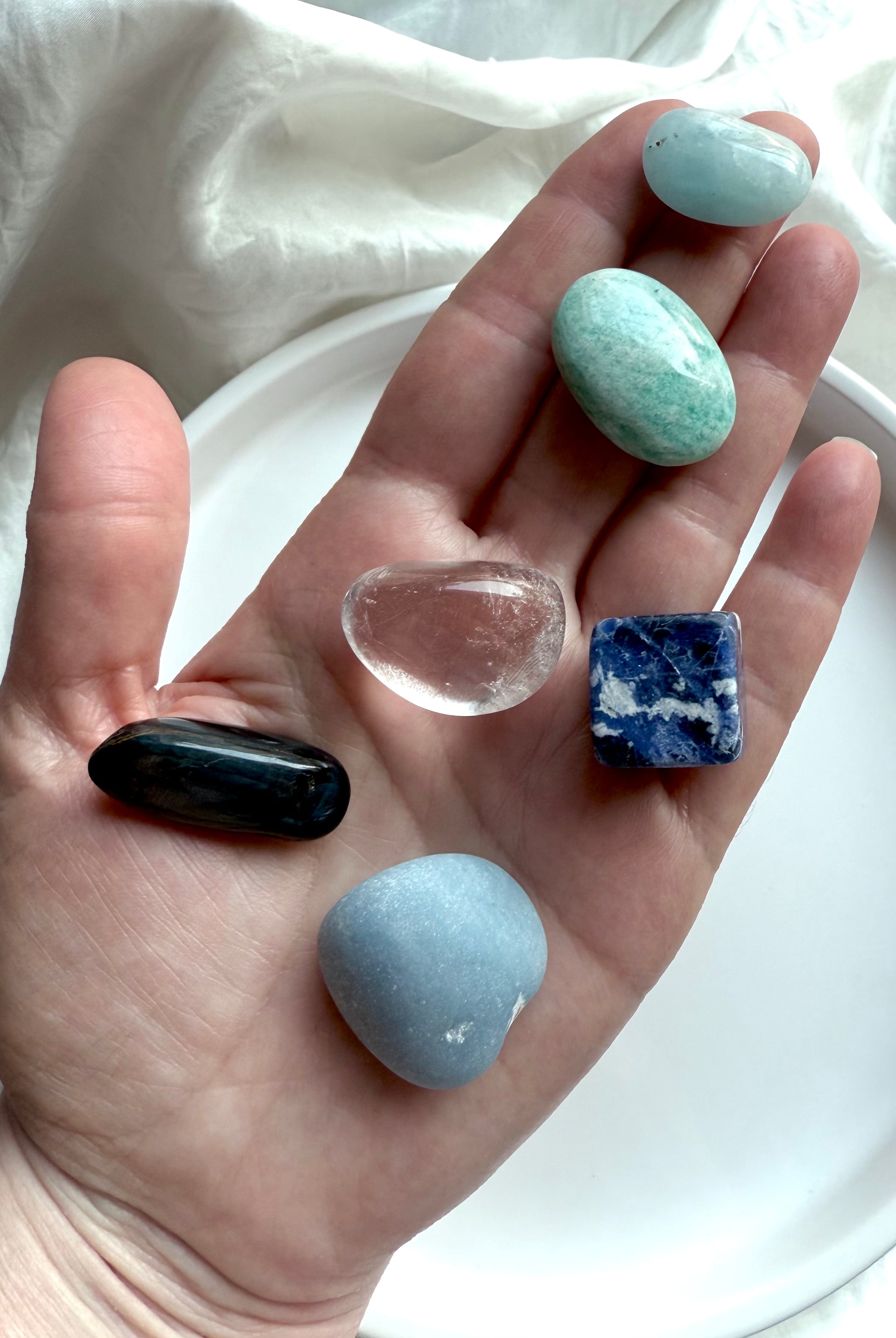 Hand holding six throat chakra crystals — Angelite, Sodalite, Amazonite, Aquamarine, Blue Tiger’s Eye, and Clear Quartz