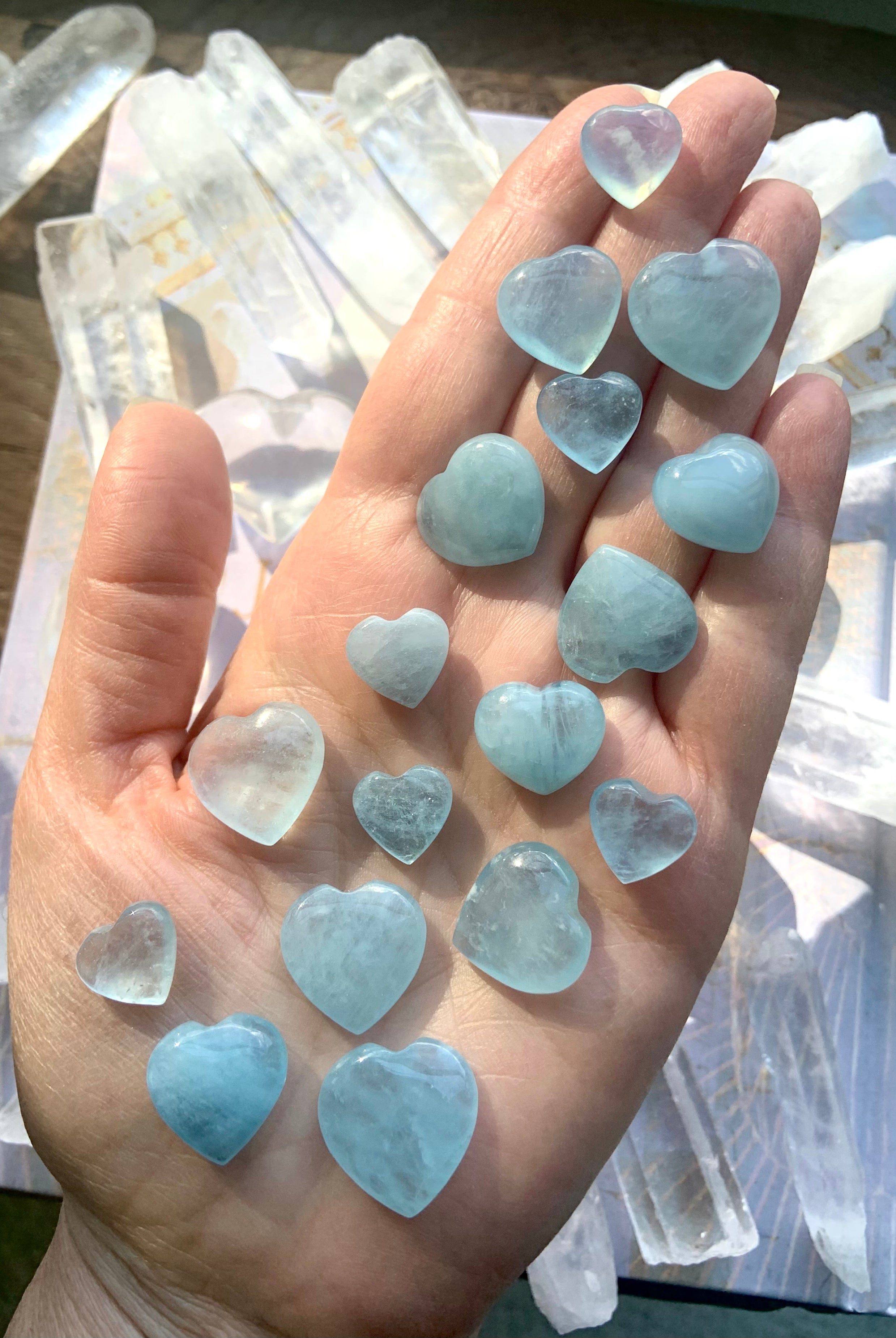 Aquamarine hearts displayed on a hand above a tabletop with quartz points — healing crystals for peace, emotional balance + open communication.