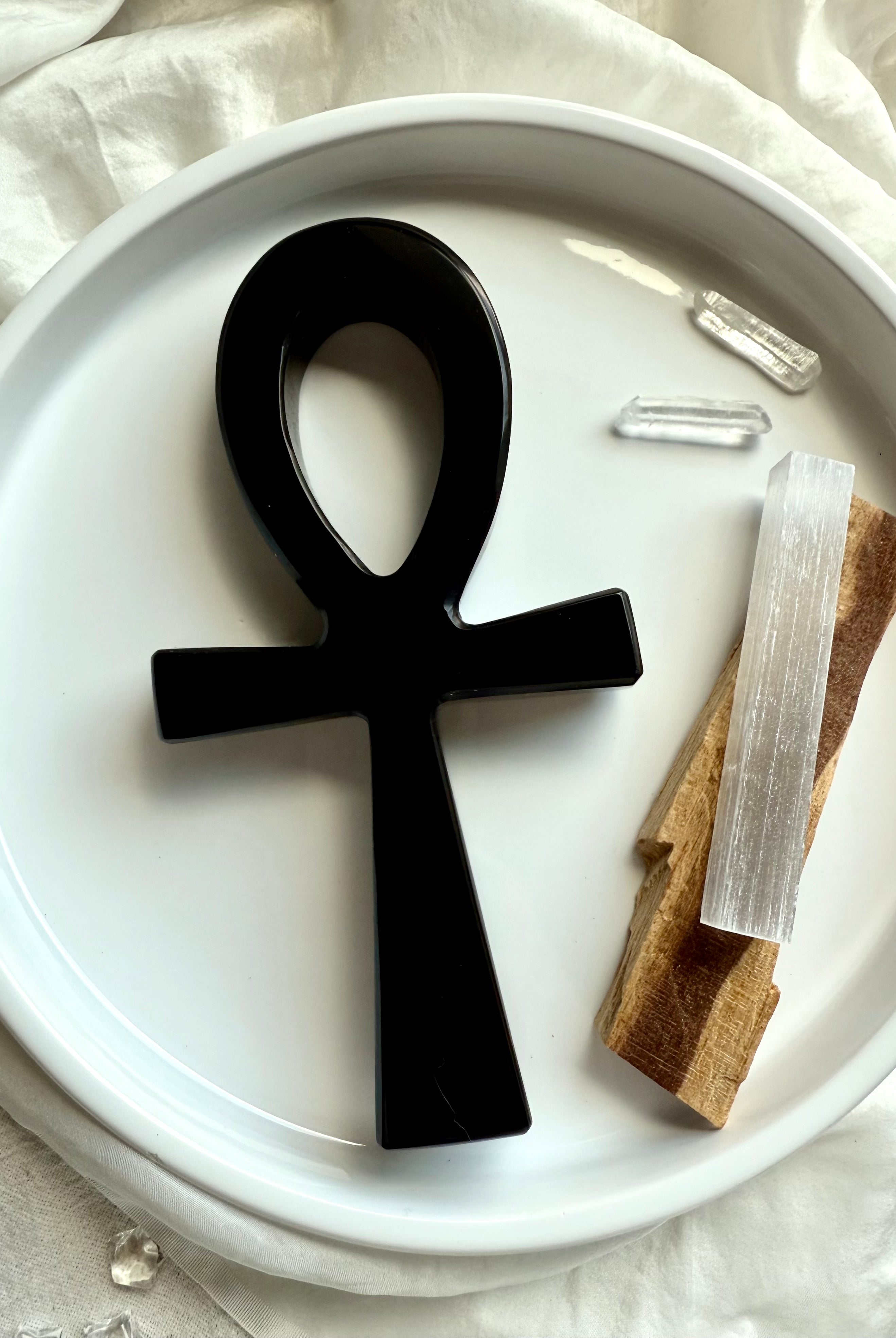 Flatlay of a Black ankh symbol on a white plate with crystals and palo santo sticks.