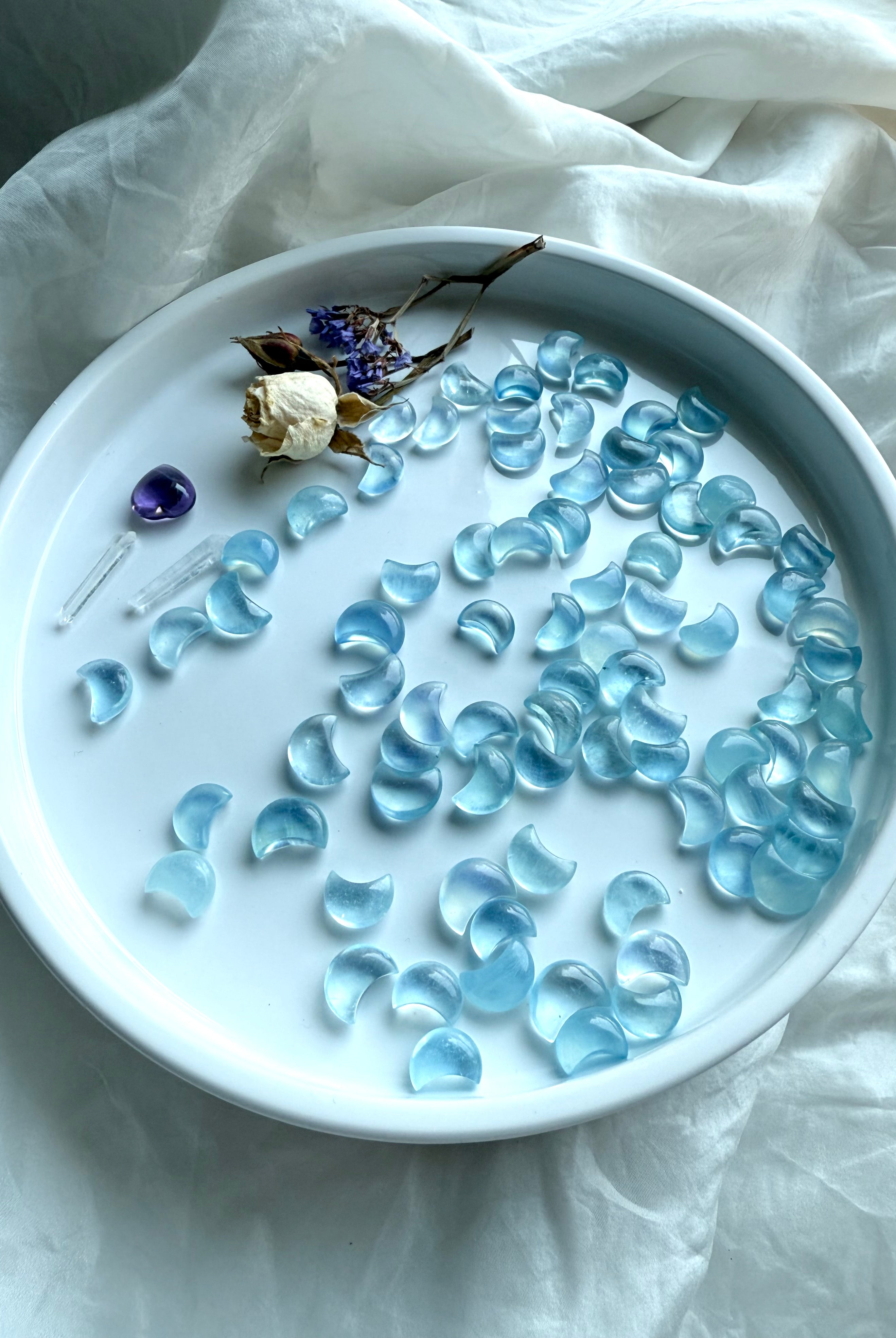 Aquamarine Mini Moons displayed in a wide array on a round white tray with dried rose, purple statice, and a tiny amethyst heart — celestial crystal carvings in pale blue hues