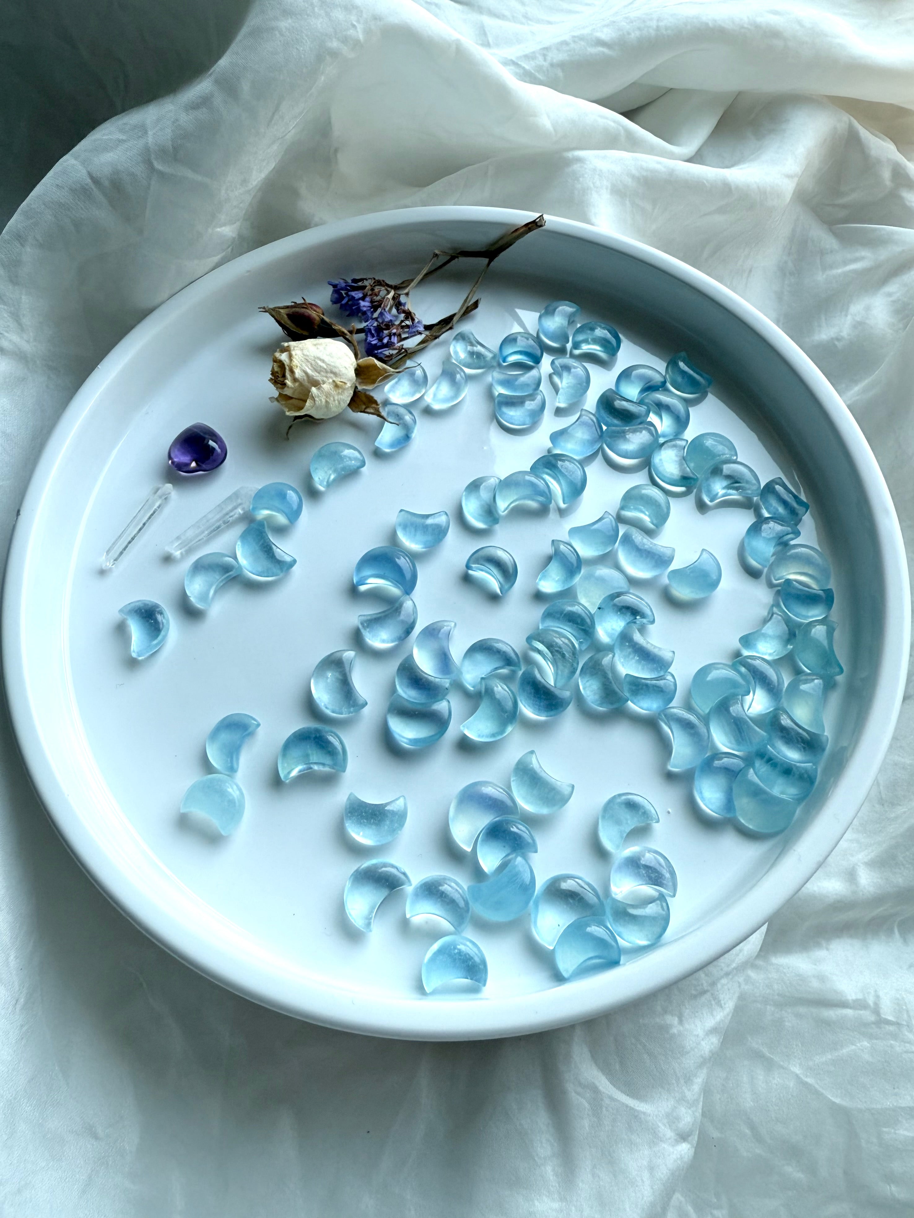 Aquamarine Mini Moons displayed in a wide array on a round white tray with dried rose, purple statice, and a tiny amethyst heart — celestial crystal carvings in pale blue hues