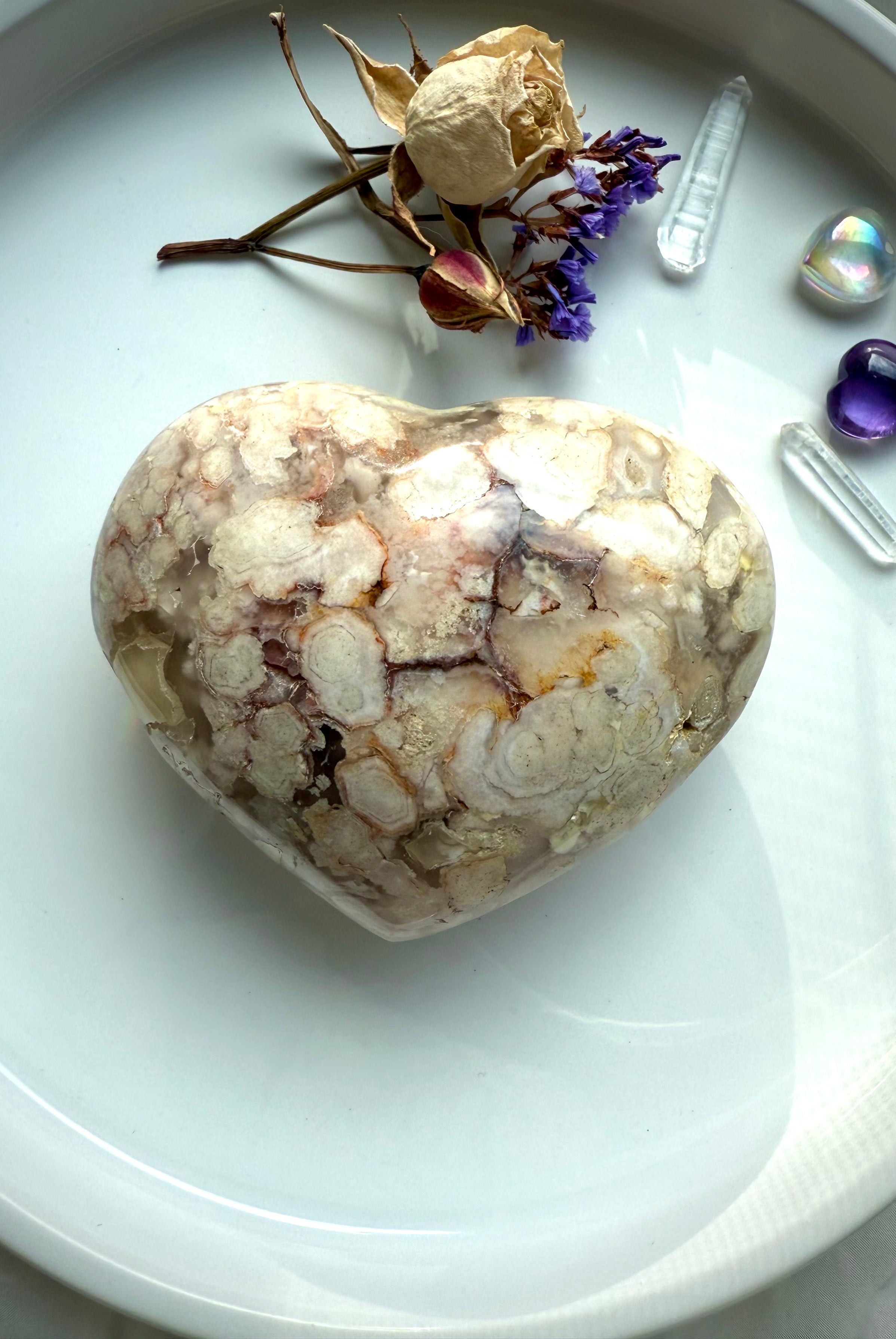 Flower Agate heart crystal with soft pink, cream, and white blossom-like patterns displayed on a dish with dried flowers — healing stone for emotional growth, comfort, and inner peace.