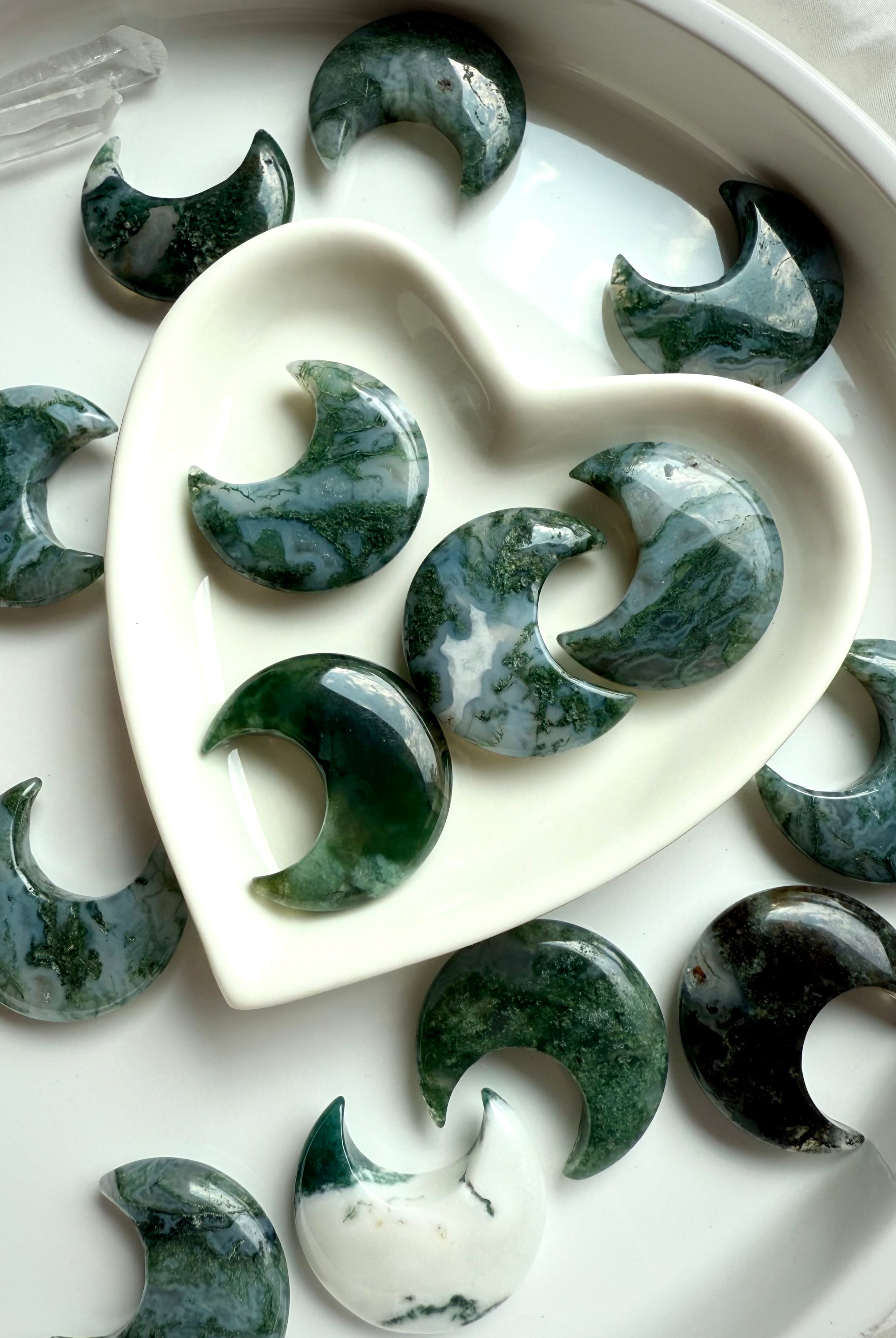 Moss Agate moon carvings in a white heart-shaped bowl with additional moons surrounding it, styled for gifting or altar display.