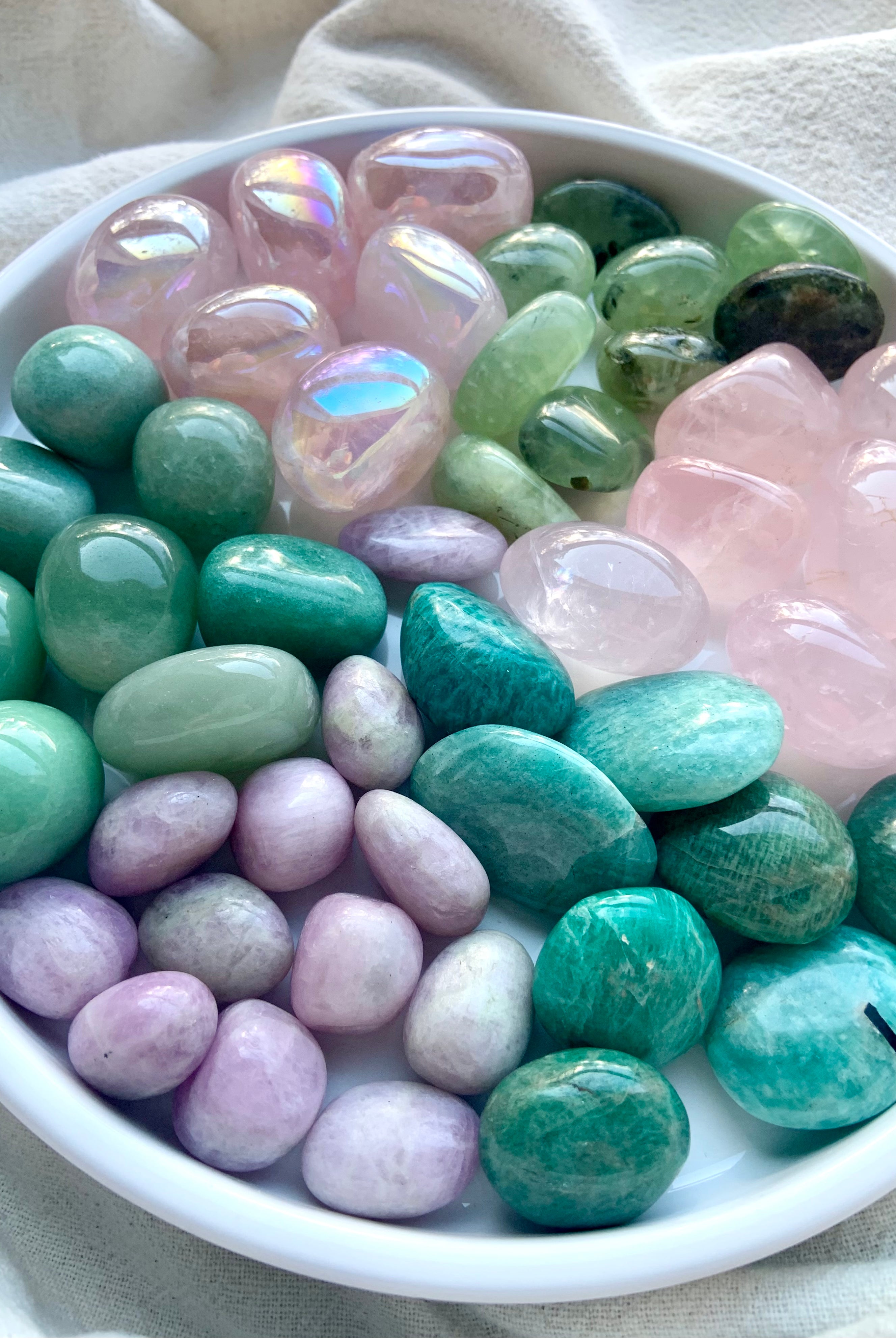 Assorted Heart Chakra healing crystals in pink and green arranged on a white plate — aura rose quartz, kunzite, amazonite, prehnite, rose quartz, and green aventurine.