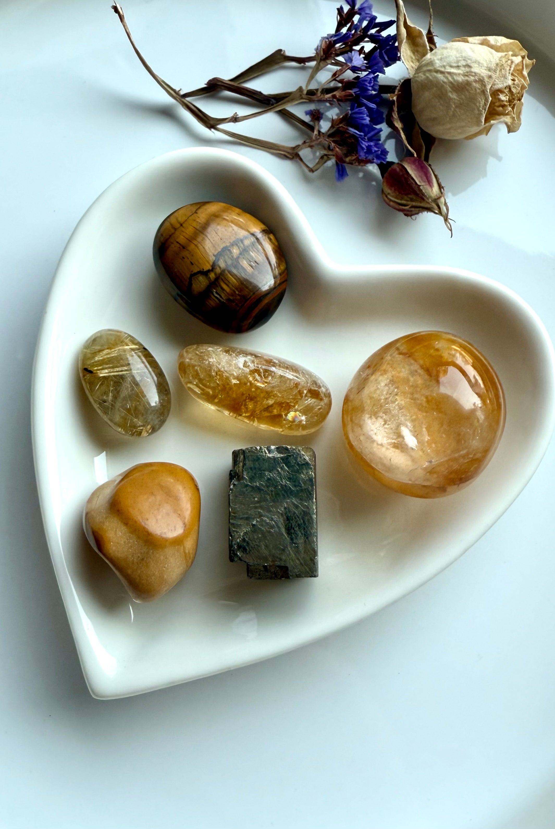 Solar Plexus Chakra crystal set arranged in a white heart-shaped bowl with dried flowers — golden healer, citrine, golden rutile, tiger’s eye, yellow mookaite, and raw pyrite