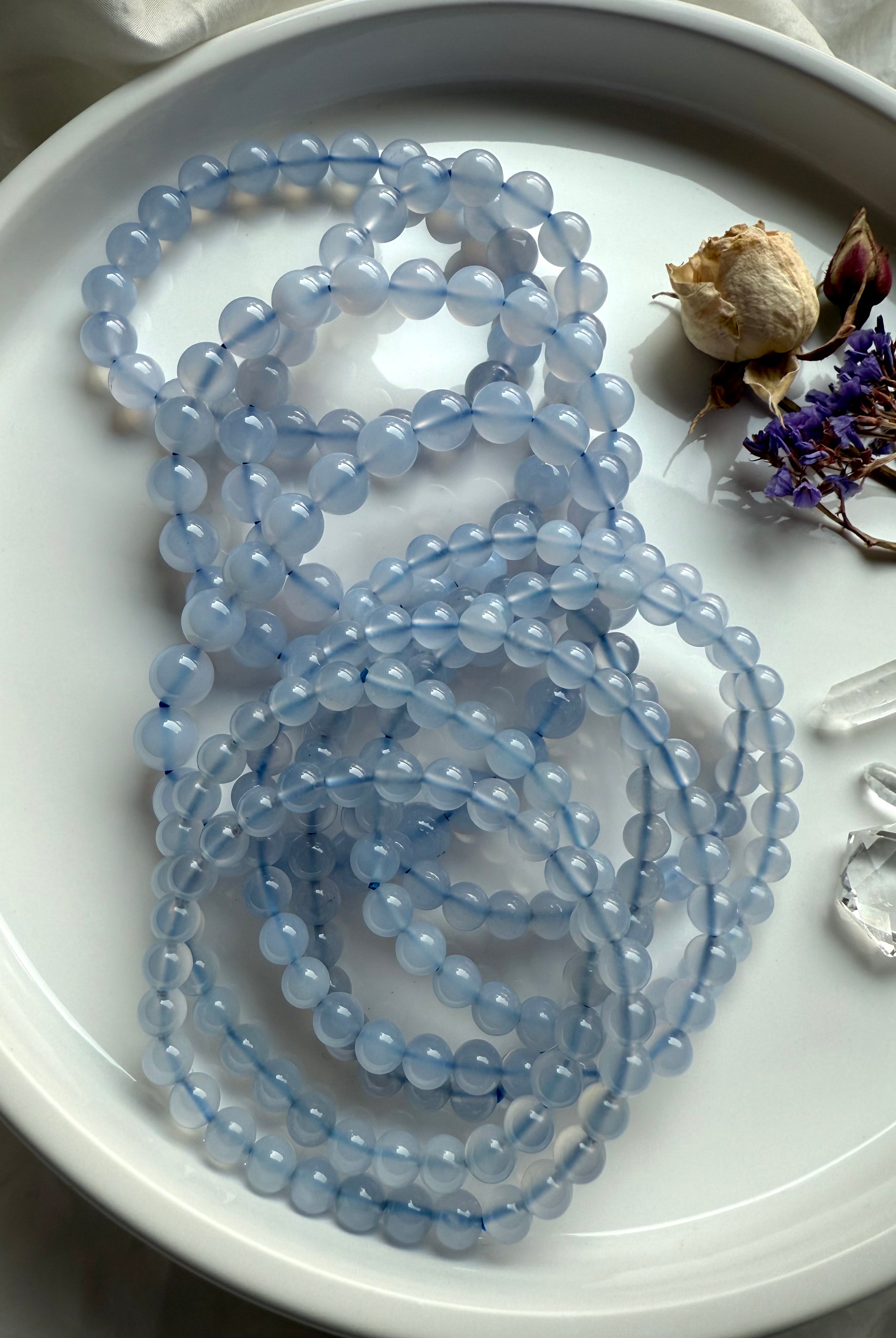 Glossy Blue Chalcedony bracelet strands with dried roses, statice flowers, and clear quartz points on white dish