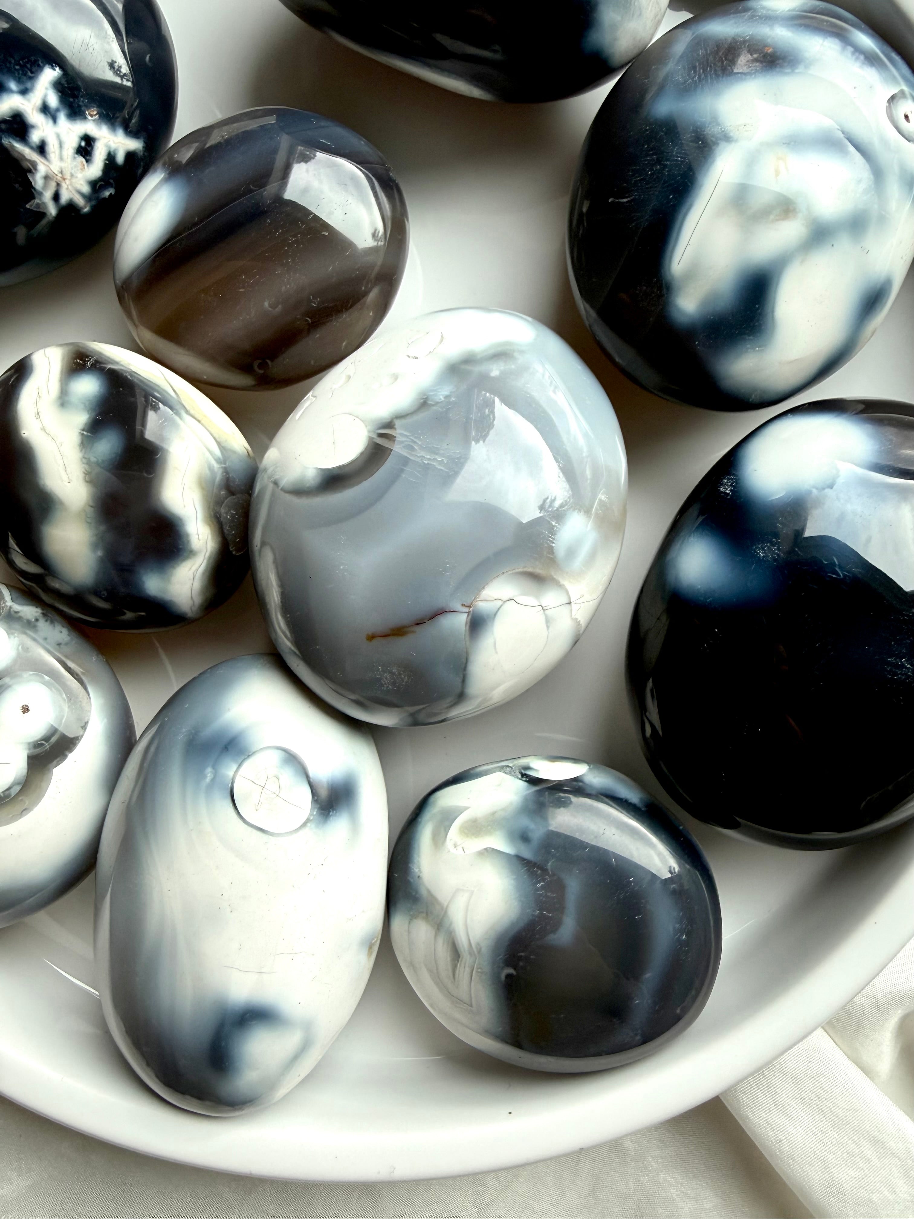 Close-up of Orca Agate palm stones resting in a white tray, showing smooth polish and natural gray, black, and white agate formations