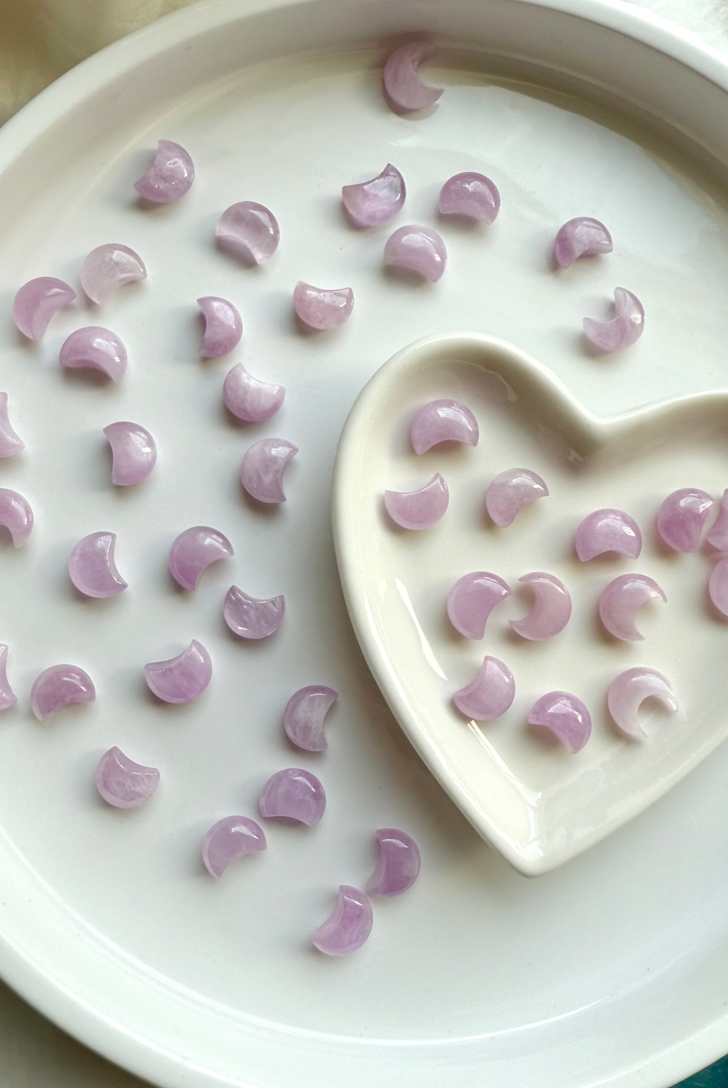 Pink Kunzite crescent moon carvings in a heart-shaped bowl with more moons scattered on a white plate — soothing crystals for heart chakra healing.