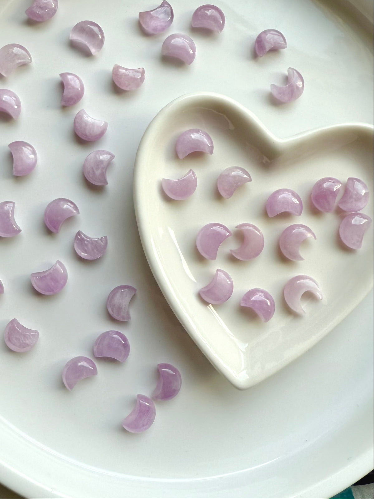 Pink Kunzite crescent moon carvings in a heart-shaped bowl with more moons scattered on a white plate — soothing crystals for heart chakra healing.