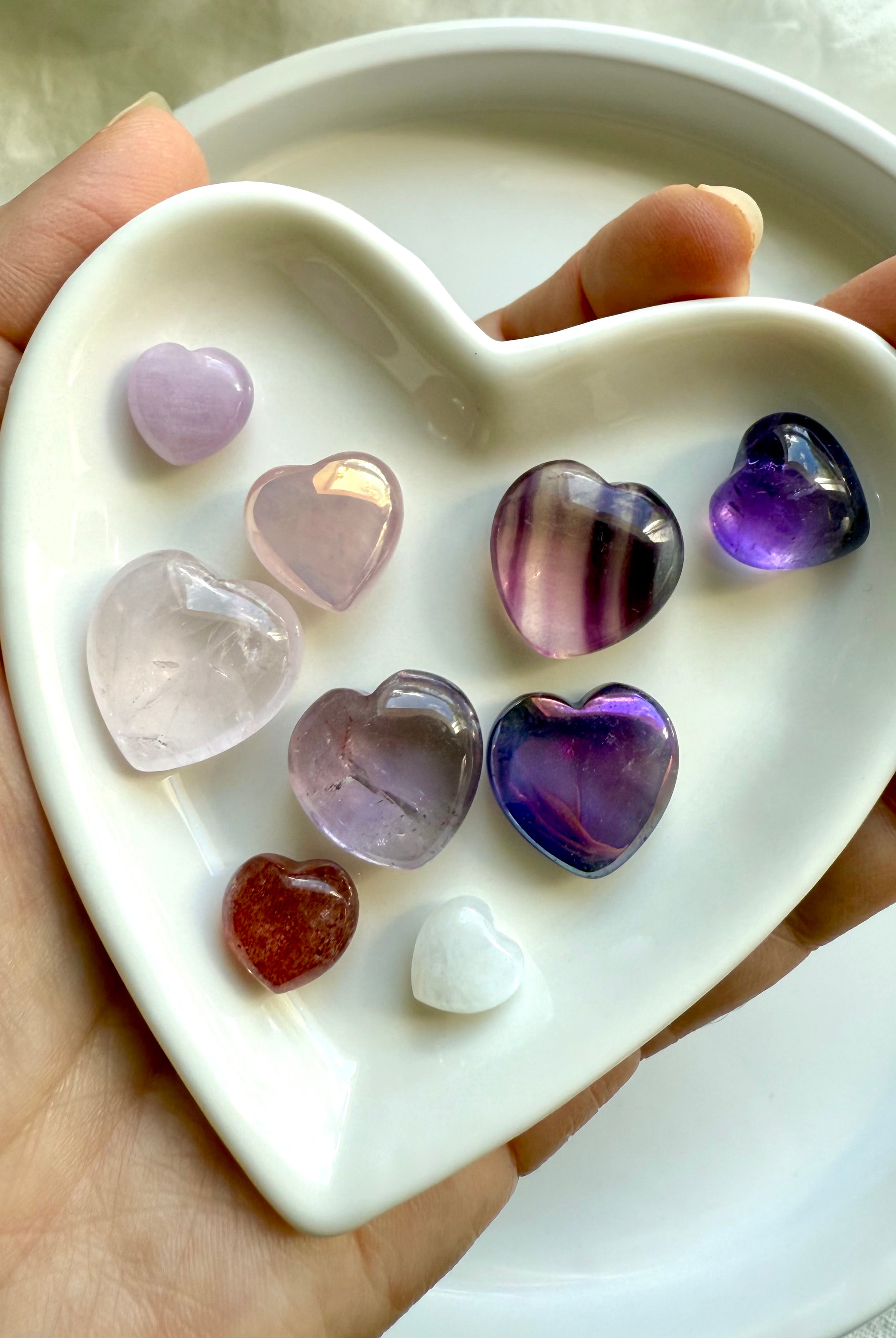 Close-up of white heart-shaped bowl holding nine pink and purple crystal hearts, premium mini gemstone carvings, held in hand.